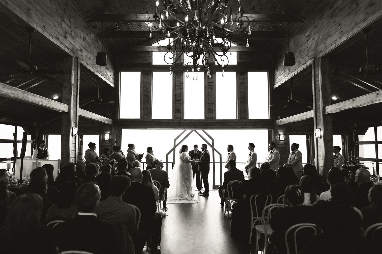 Black and white photo of wedding ceremony in rustic barn venue with chandelier and large windows.