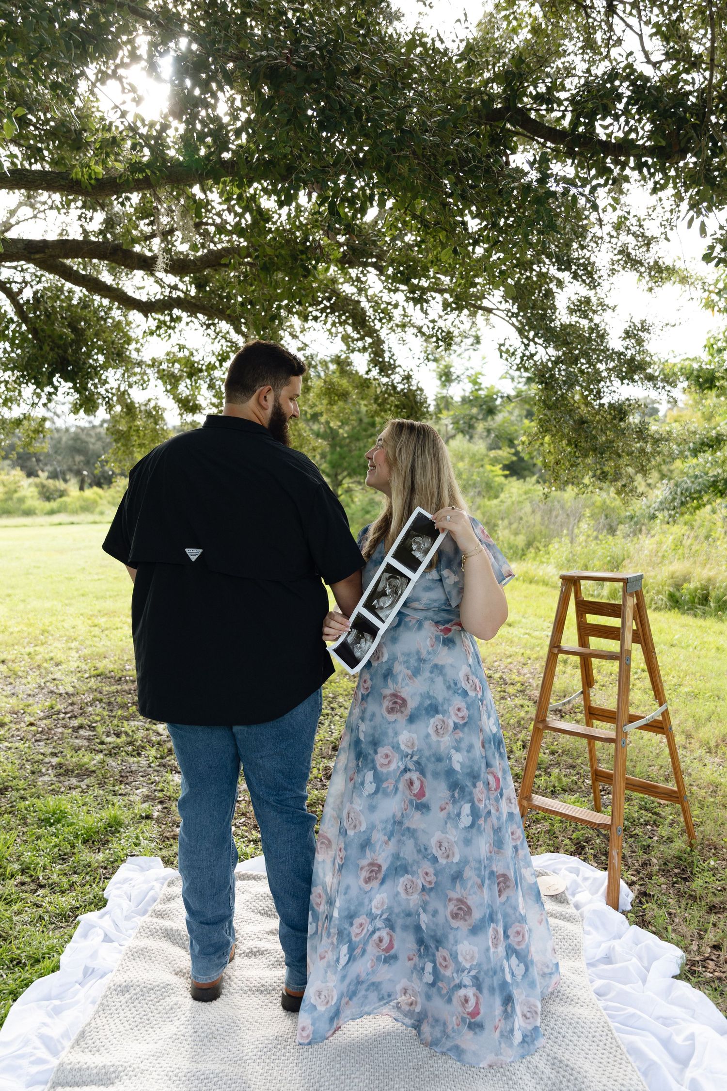Couple standing outdoors under a tree on a white blanket, holding an ultrasound photo, announcing their pregnancy.