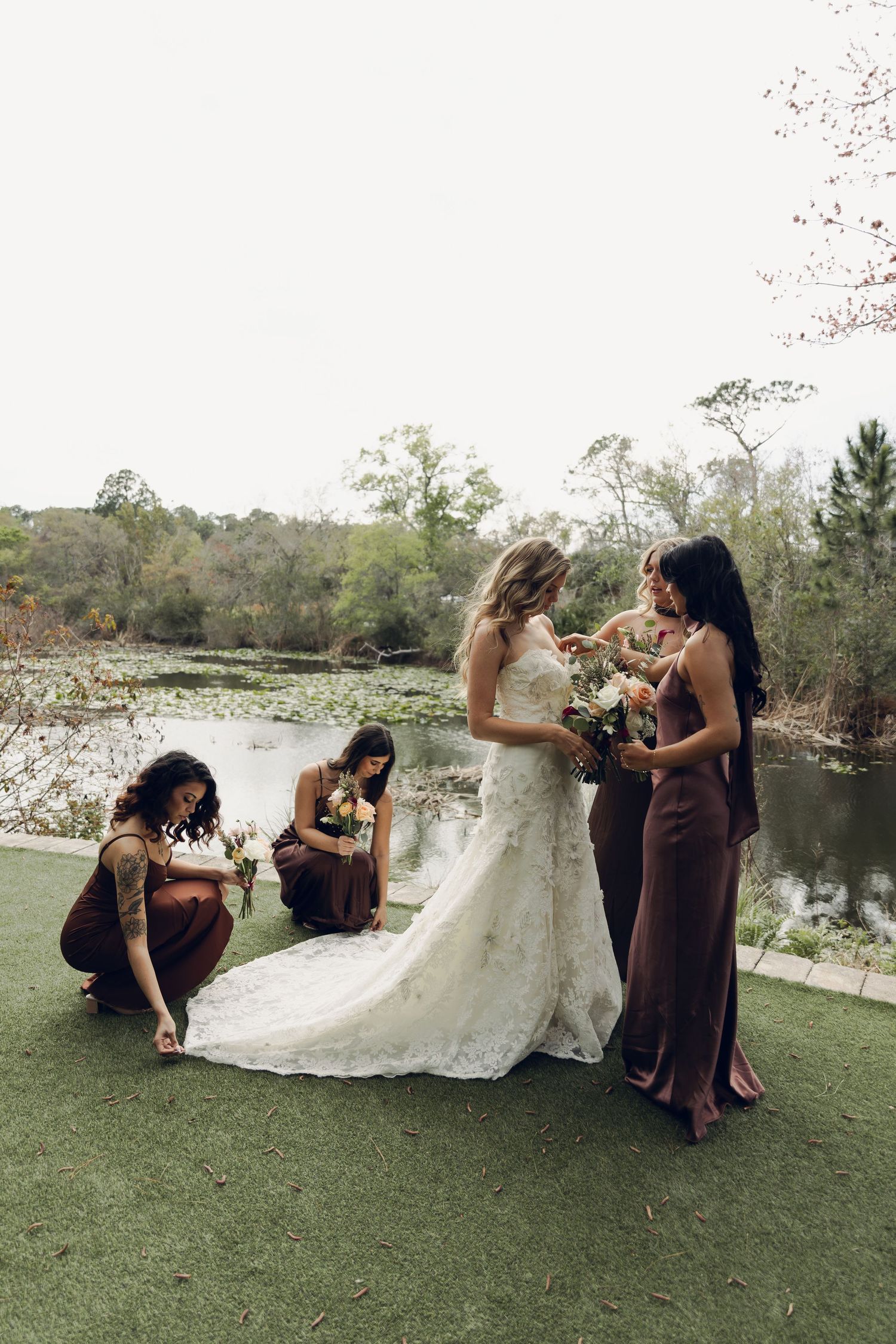 Bridesmaids in brown dresses help bride with lace train near serene pond.
