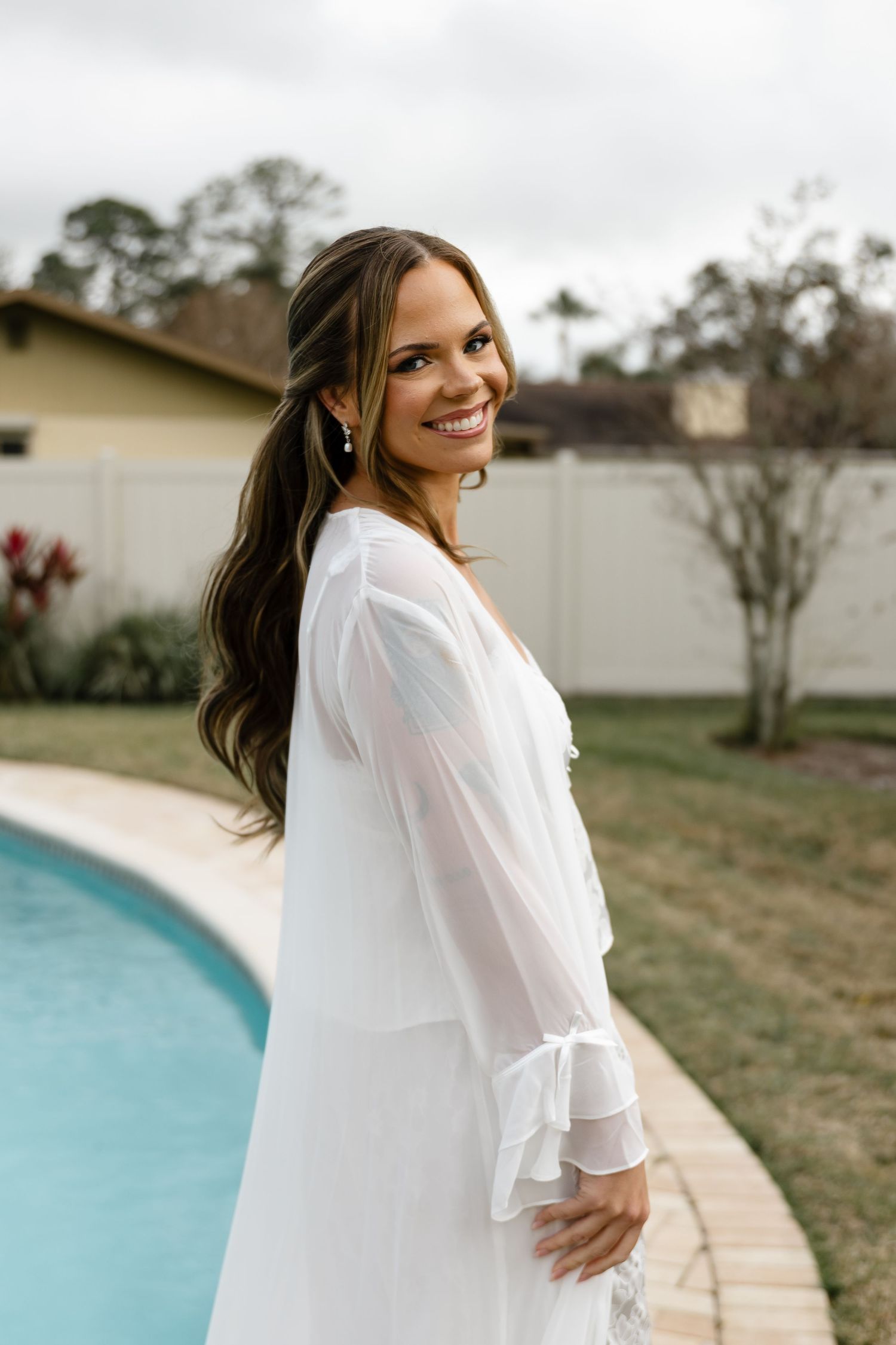 Smiling bride in a white chiffon dress posing by a pool on her wedding day.