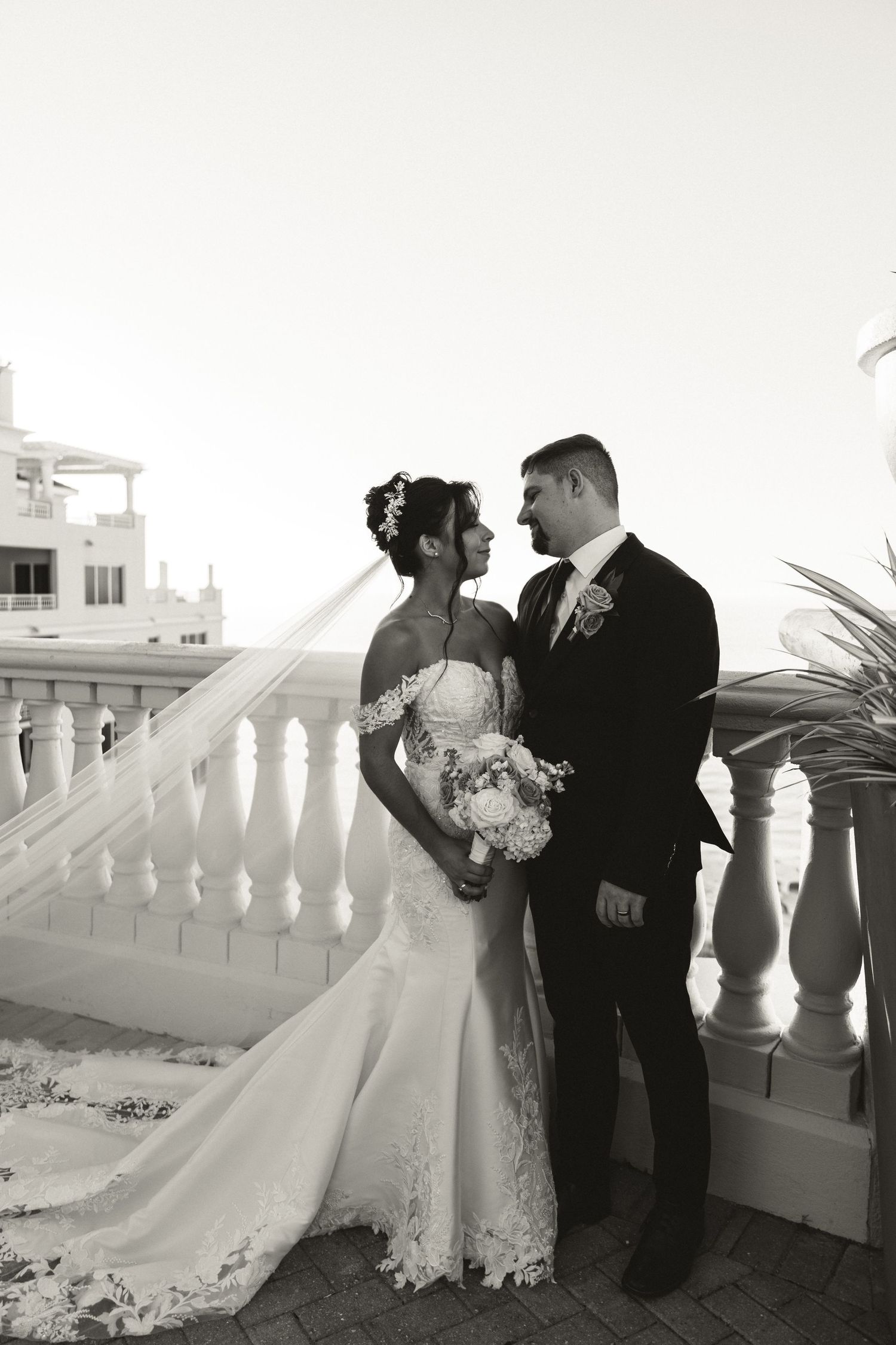 Black and white portrait of bride and groom sharing a tender moment on a seaside balcony.