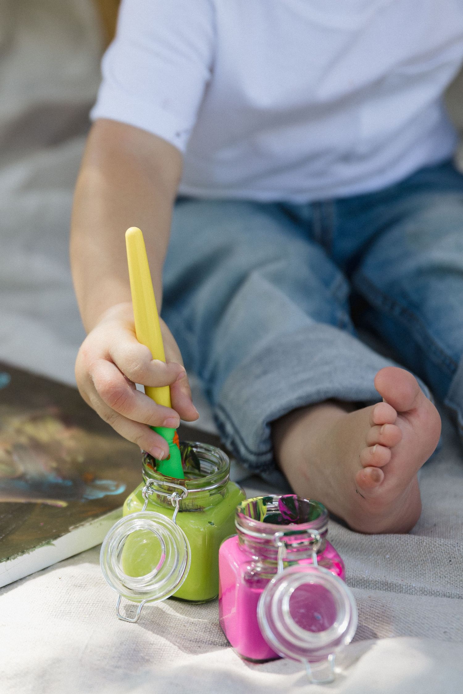 Young child sitting barefoot dipping paintbrush into green paint jar with colorful paints nearby.