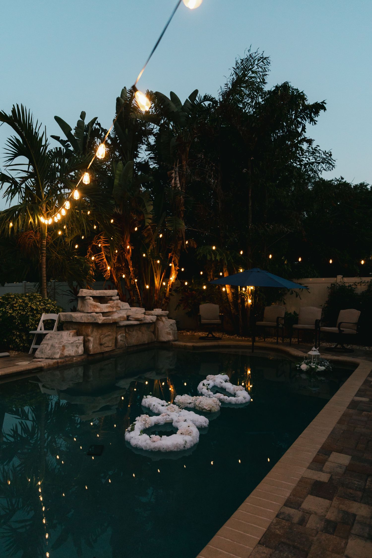 Romantic poolside wedding decor with floating floral letters and string lights at dusk.