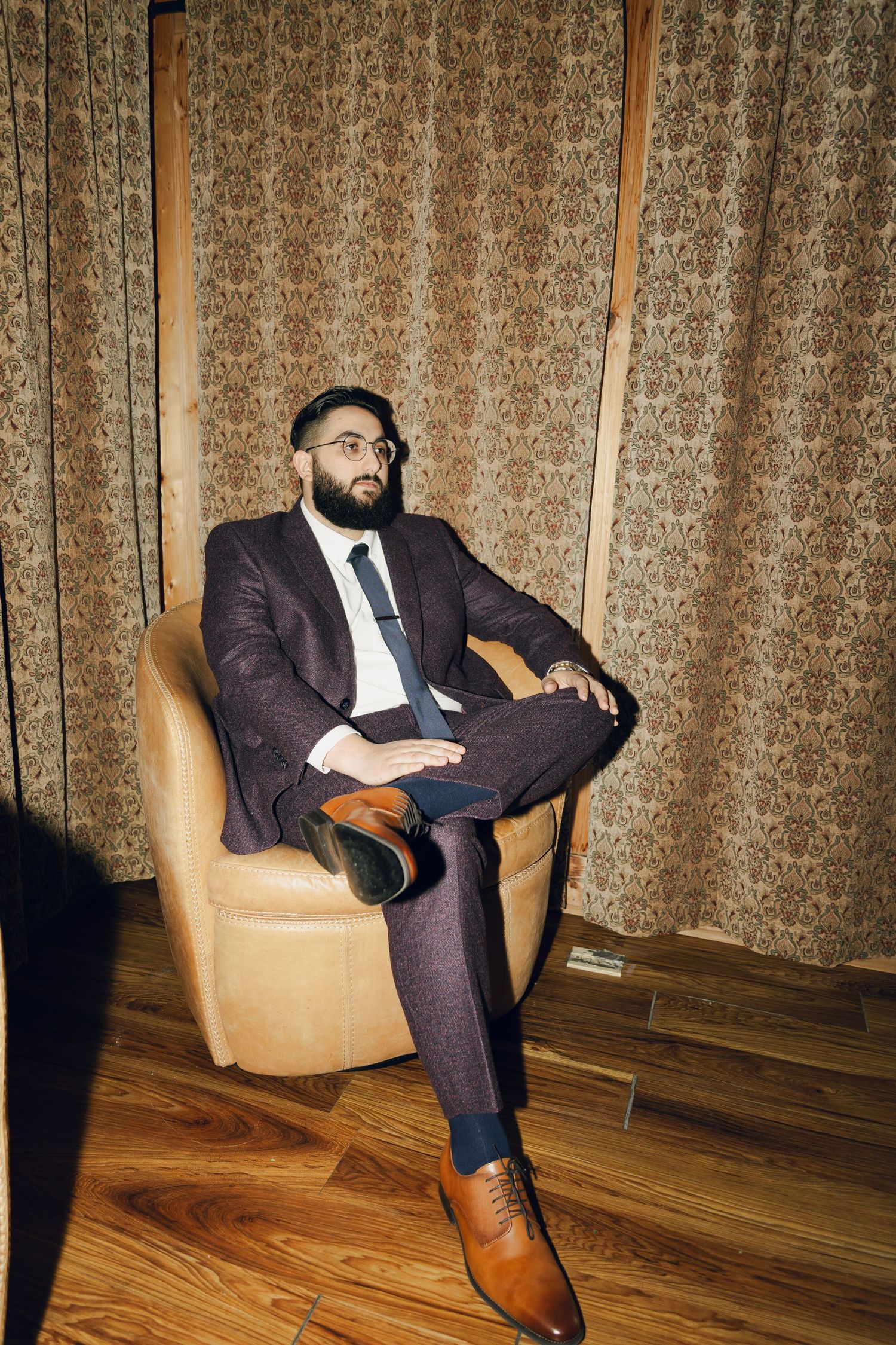 Bearded groom in dark suit and glasses sits in armchair before patterned curtains.