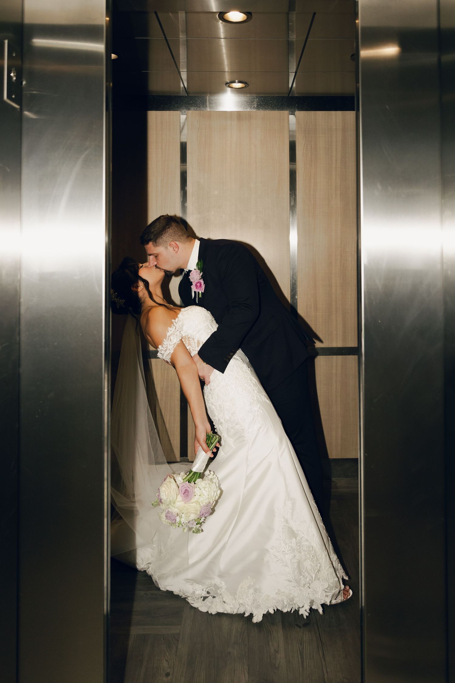 Groom dipping bride for a passionate kiss in a sleek elevator on their wedding day.