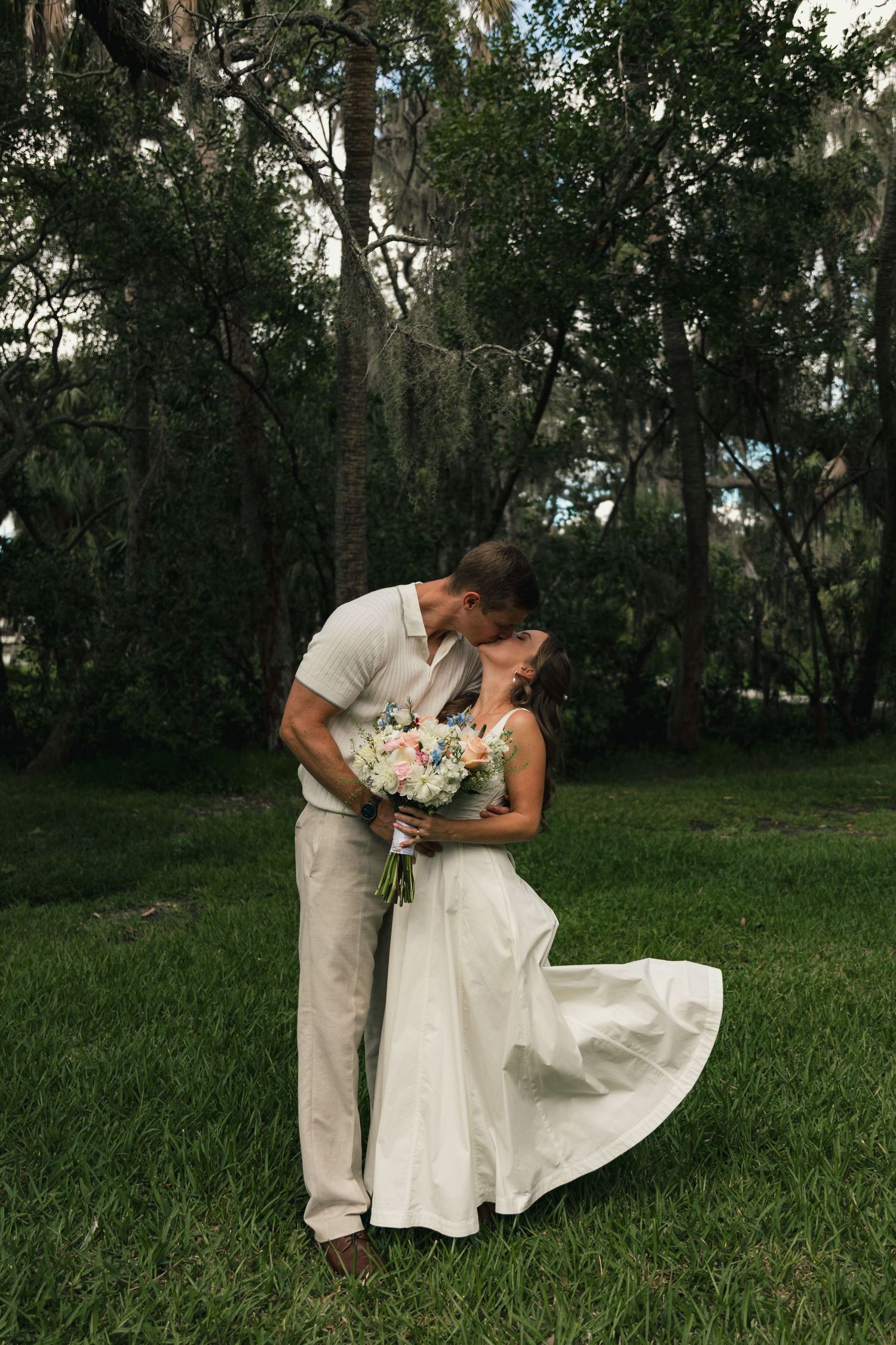 Newlywed couple sharing romantic kiss on green lawn holding pastel wedding bouquet in lush forest.