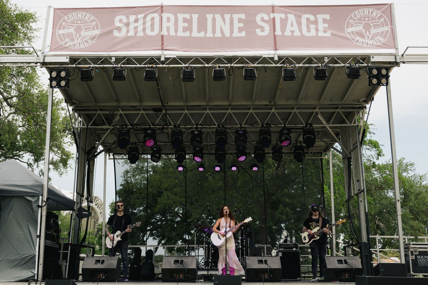 Band performing on Shoreline Stage at outdoor music festival surrounded by trees.