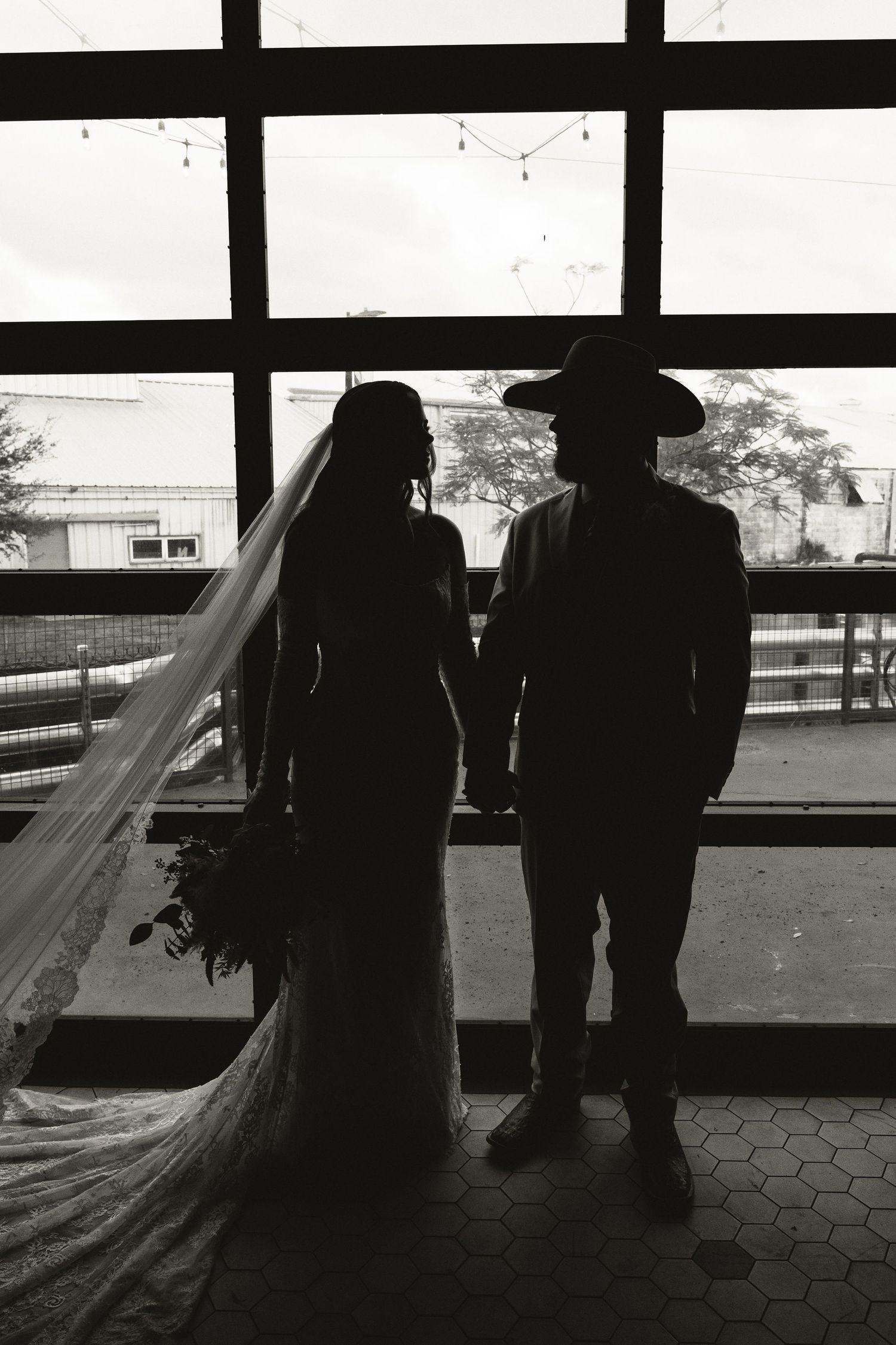 Silhouette of bride and groom holding hands in front of large windows in black and white.