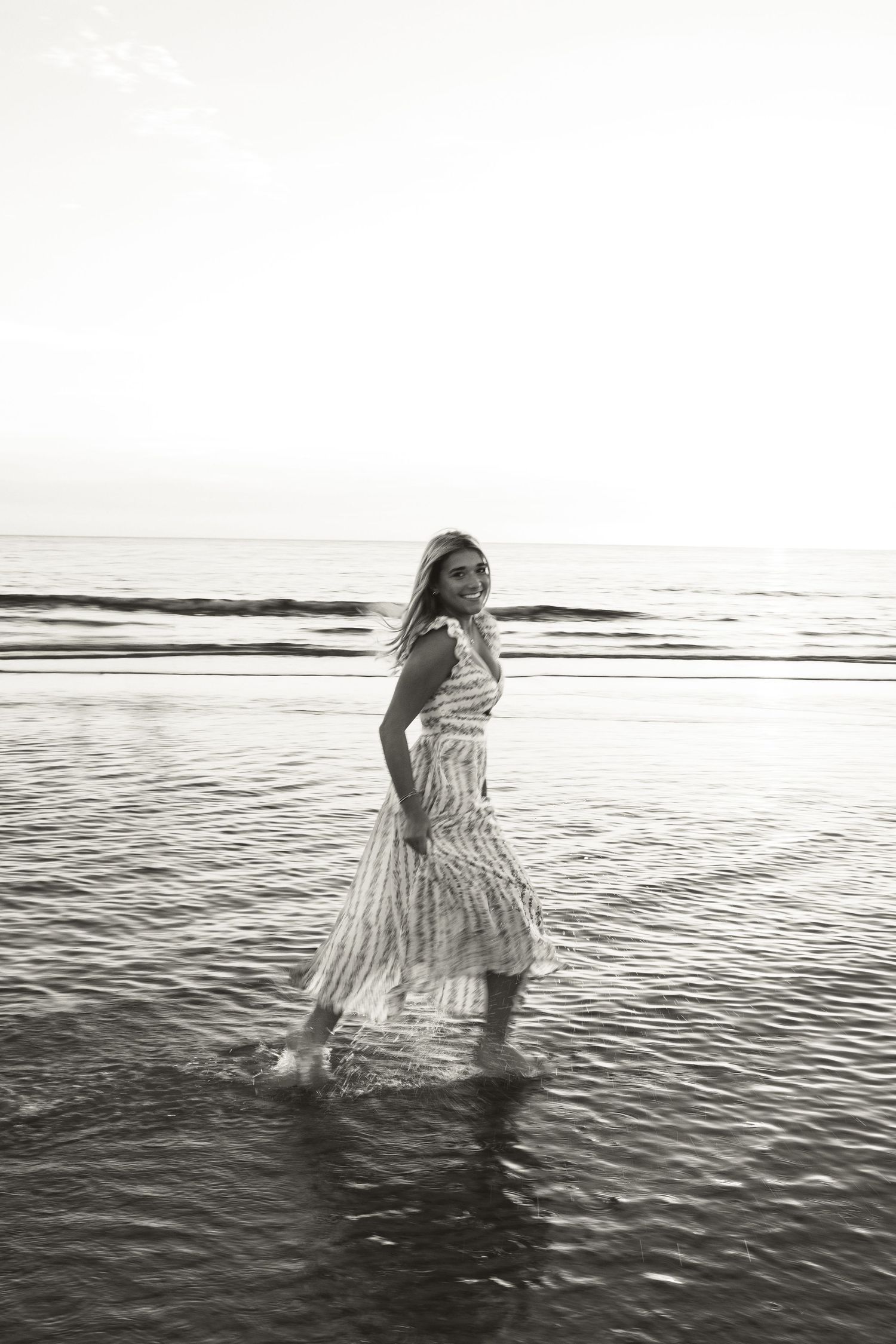 Black and white photo of a woman in a flowy dress wading through shallow ocean waves on a beach at sunset.