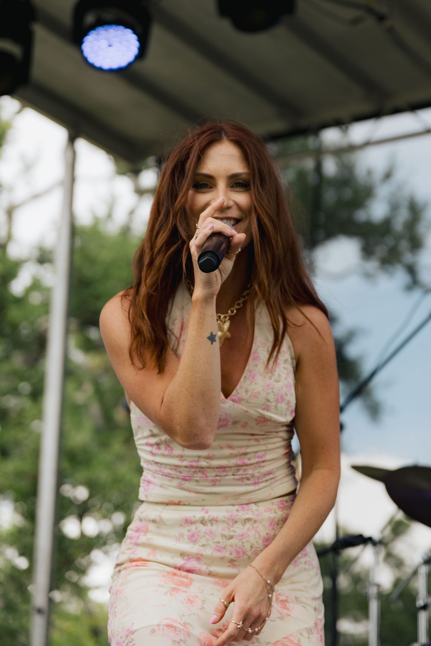 Female singer with auburn hair performing on outdoor stage in floral dress holding microphone.
