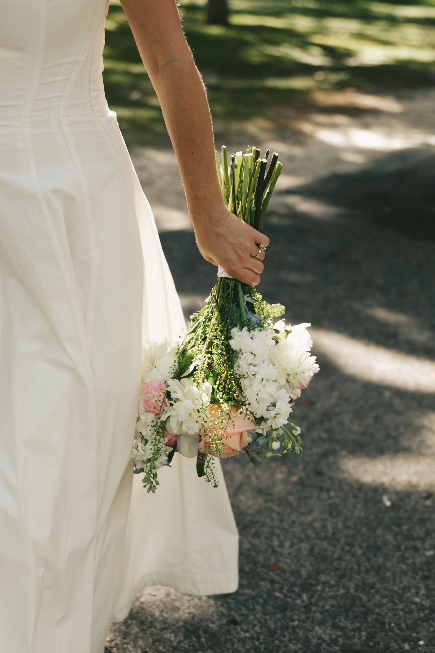 Bride in white wedding dress holding elegant pink and white floral bouquet in dappled sunlight.