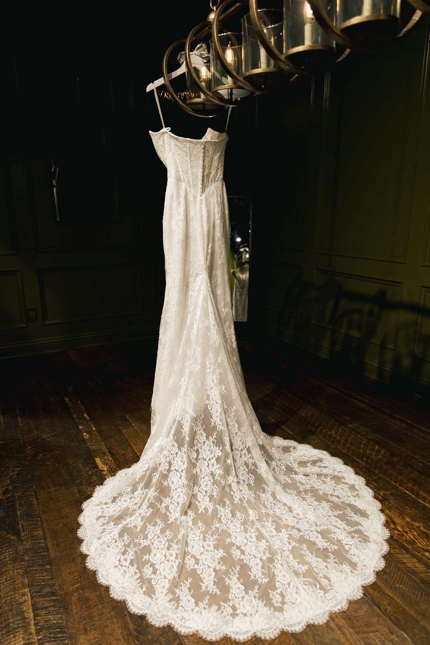 Lace wedding gown with cathedral train hanging from a rustic chandelier in a dark room.