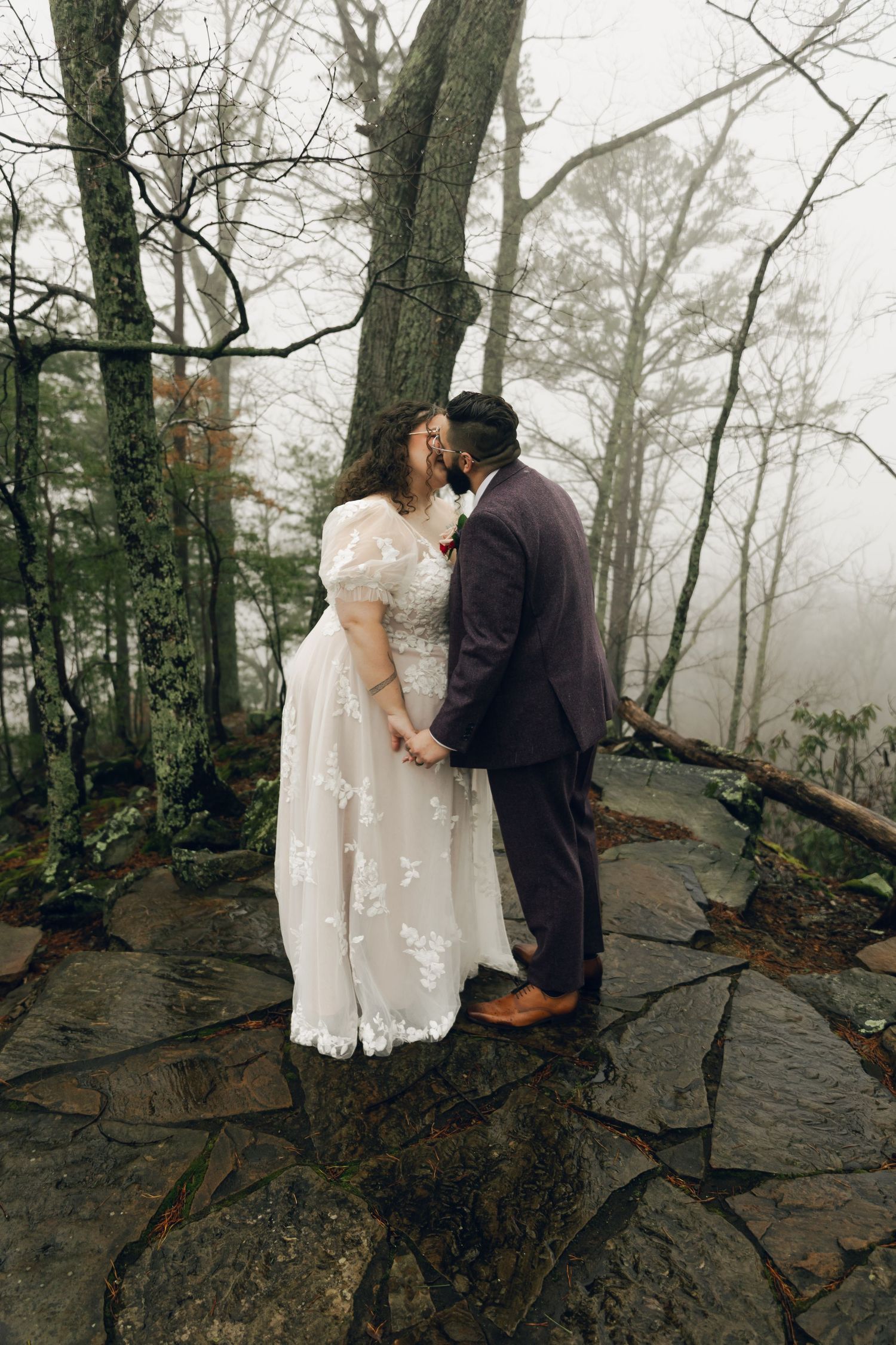 Bride and groom share a kiss on foggy forest stone path in winter.