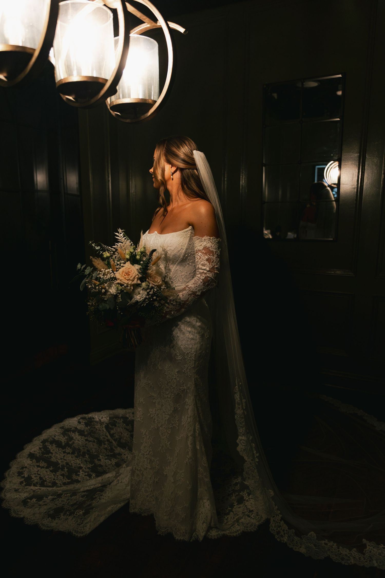 Bride in an off-shoulder lace gown with veil holding a bouquet under warm chandelier light.