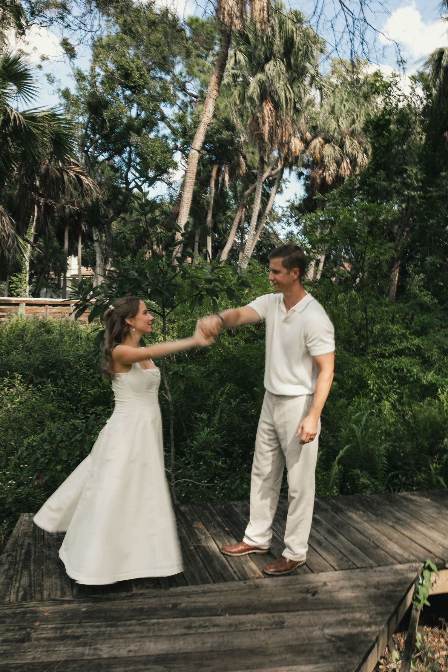 Happy bride and groom dancing together on wooden boardwalk surrounded by lush tropical greenery.