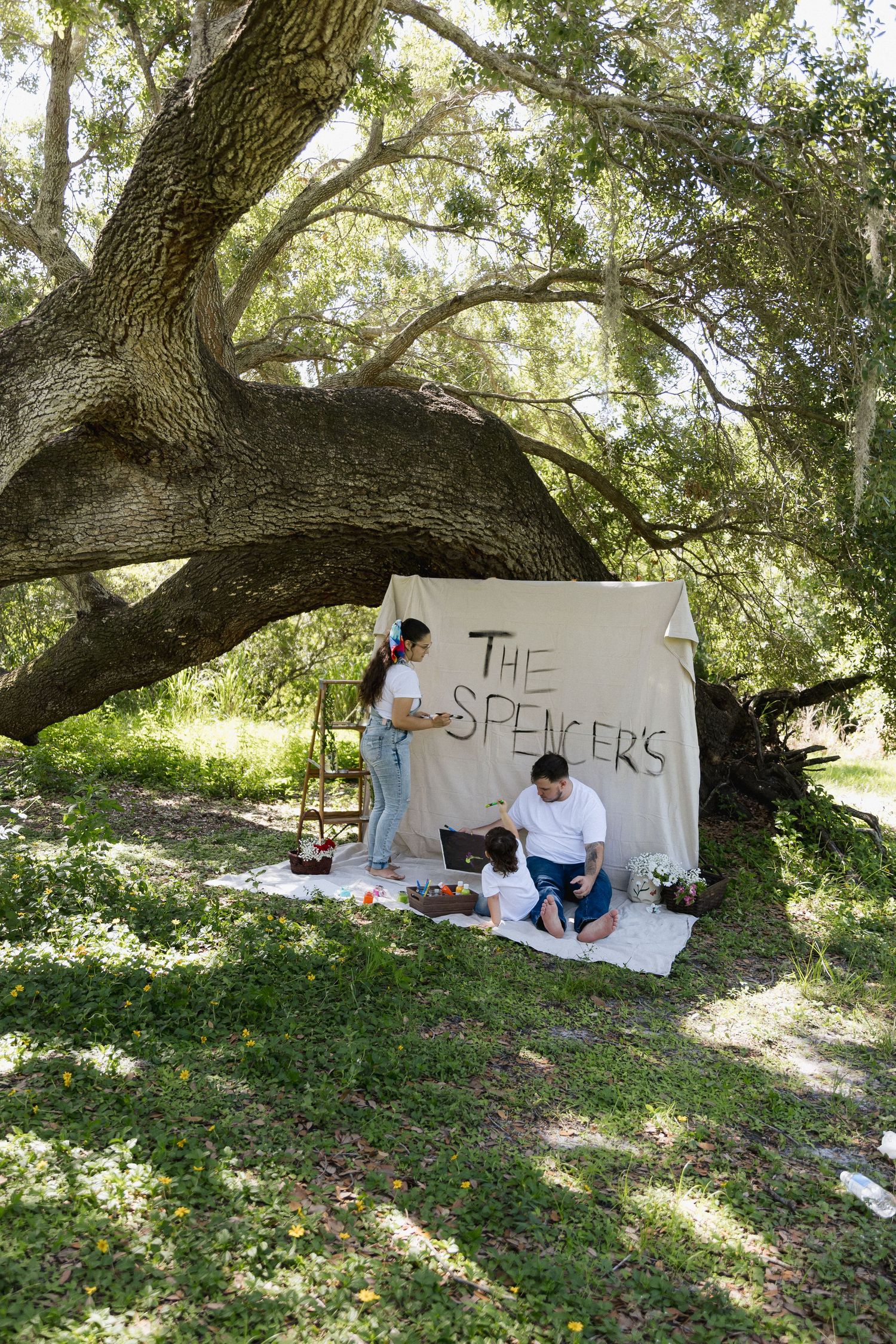 Family posing under large oak tree with handmade The Spencers banner at outdoor photo shoot.