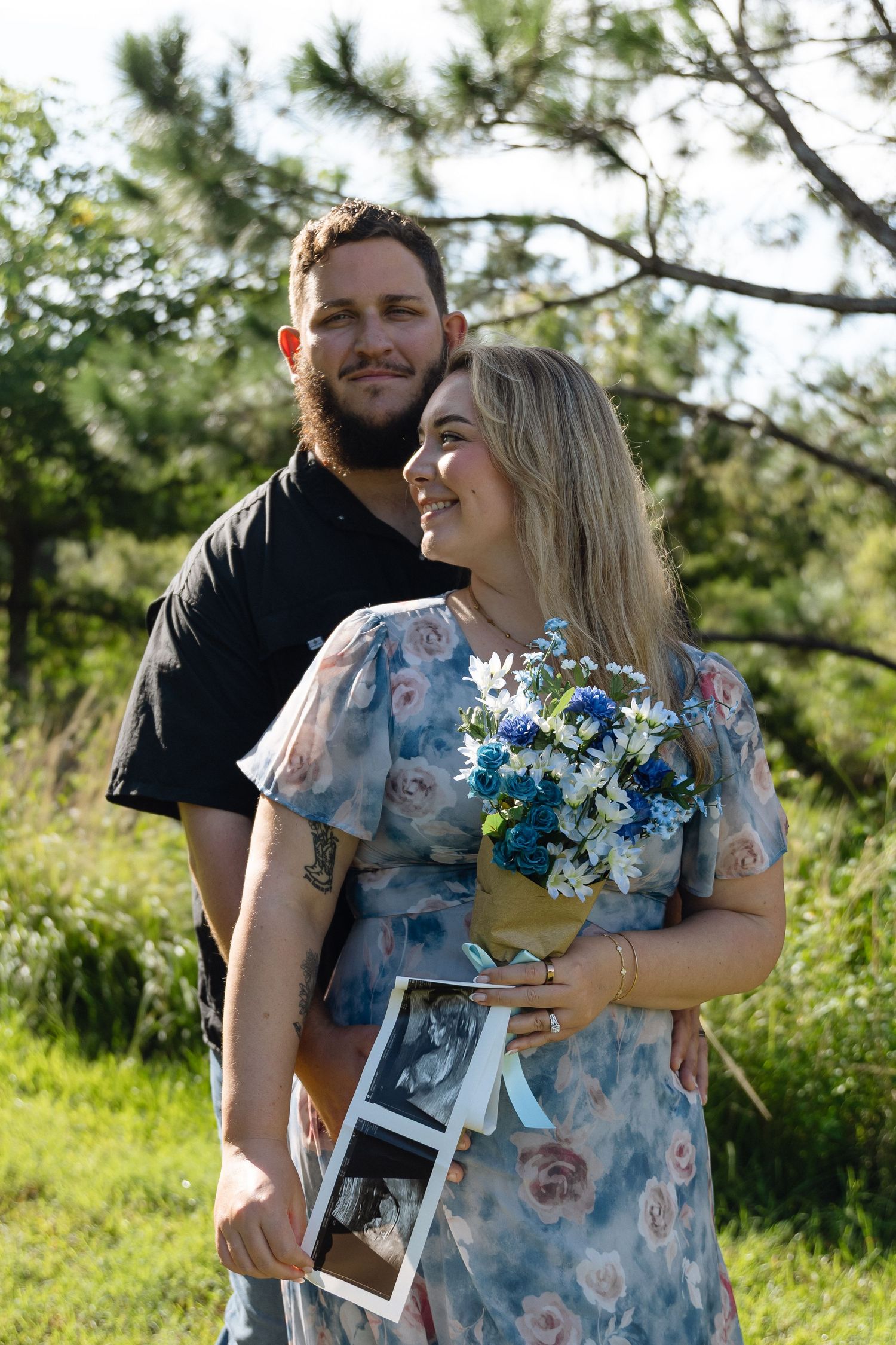 Couple outdoors holding blue and white flowers and ultrasound photos, smiling in a pregnancy announcement portrait.