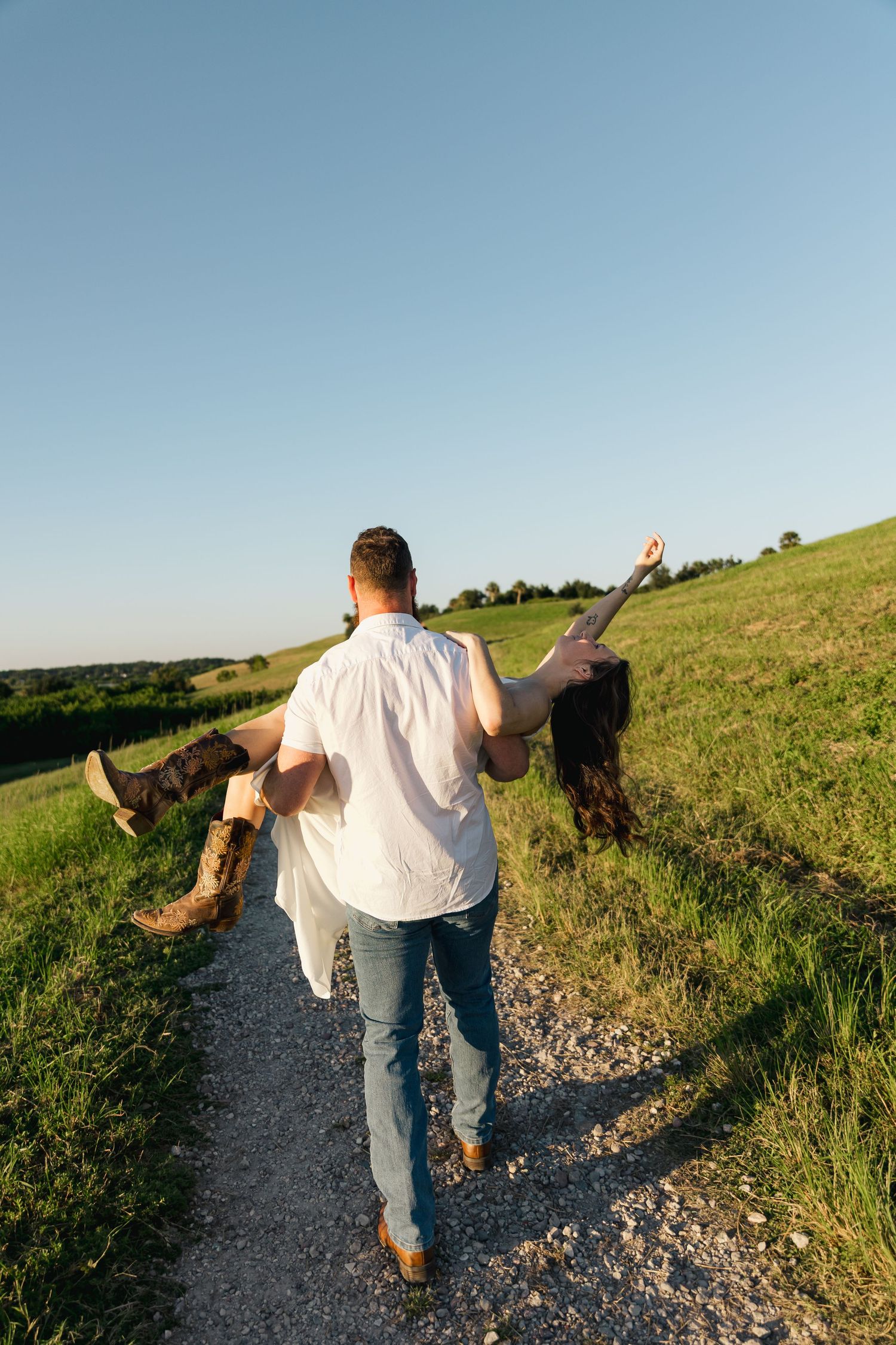 Man carrying woman in white dress along a scenic gravel path through green fields under a clear blue sky.
