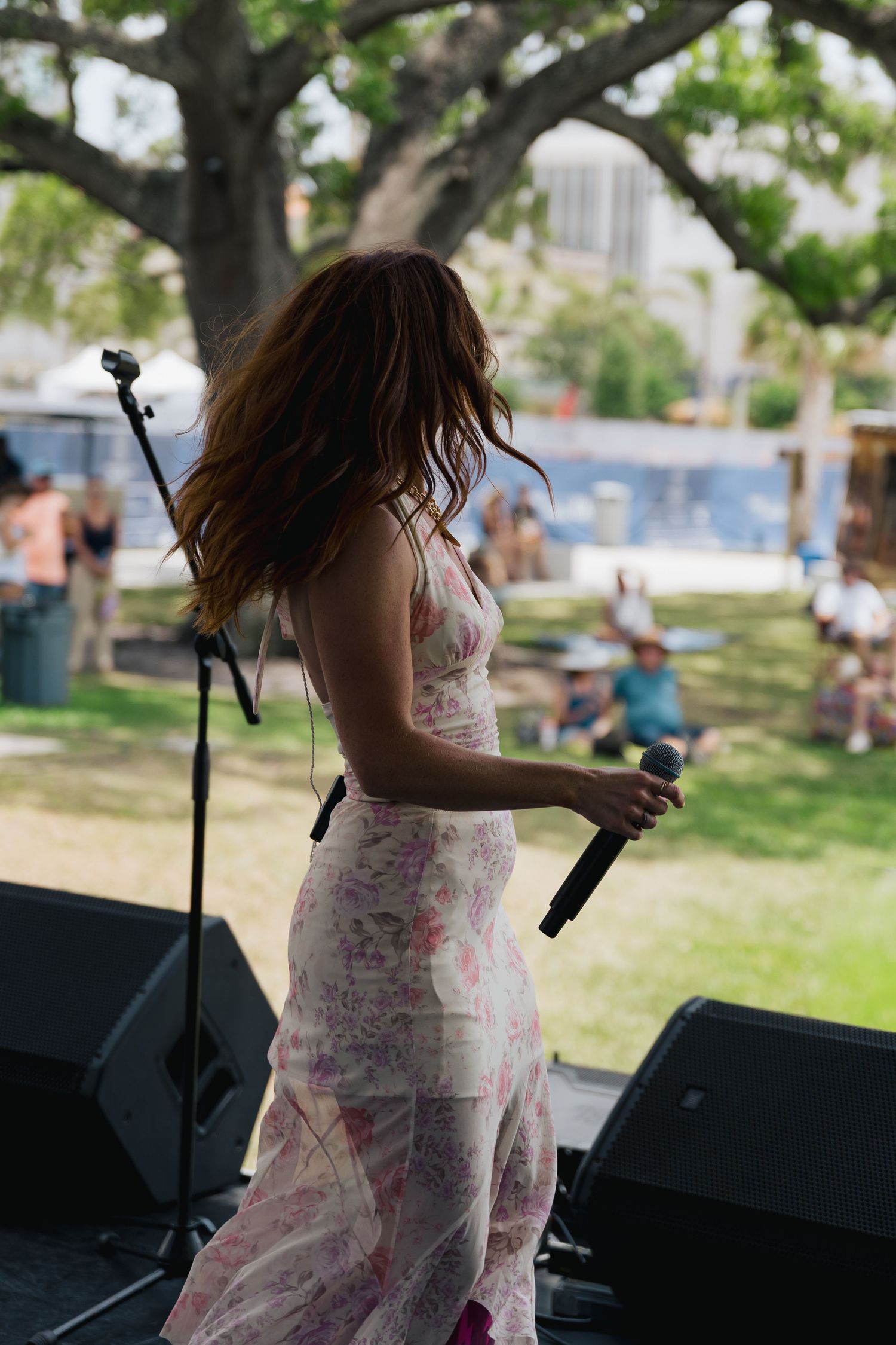Female performer in floral dress holding microphone on outdoor stage with crowd in background.