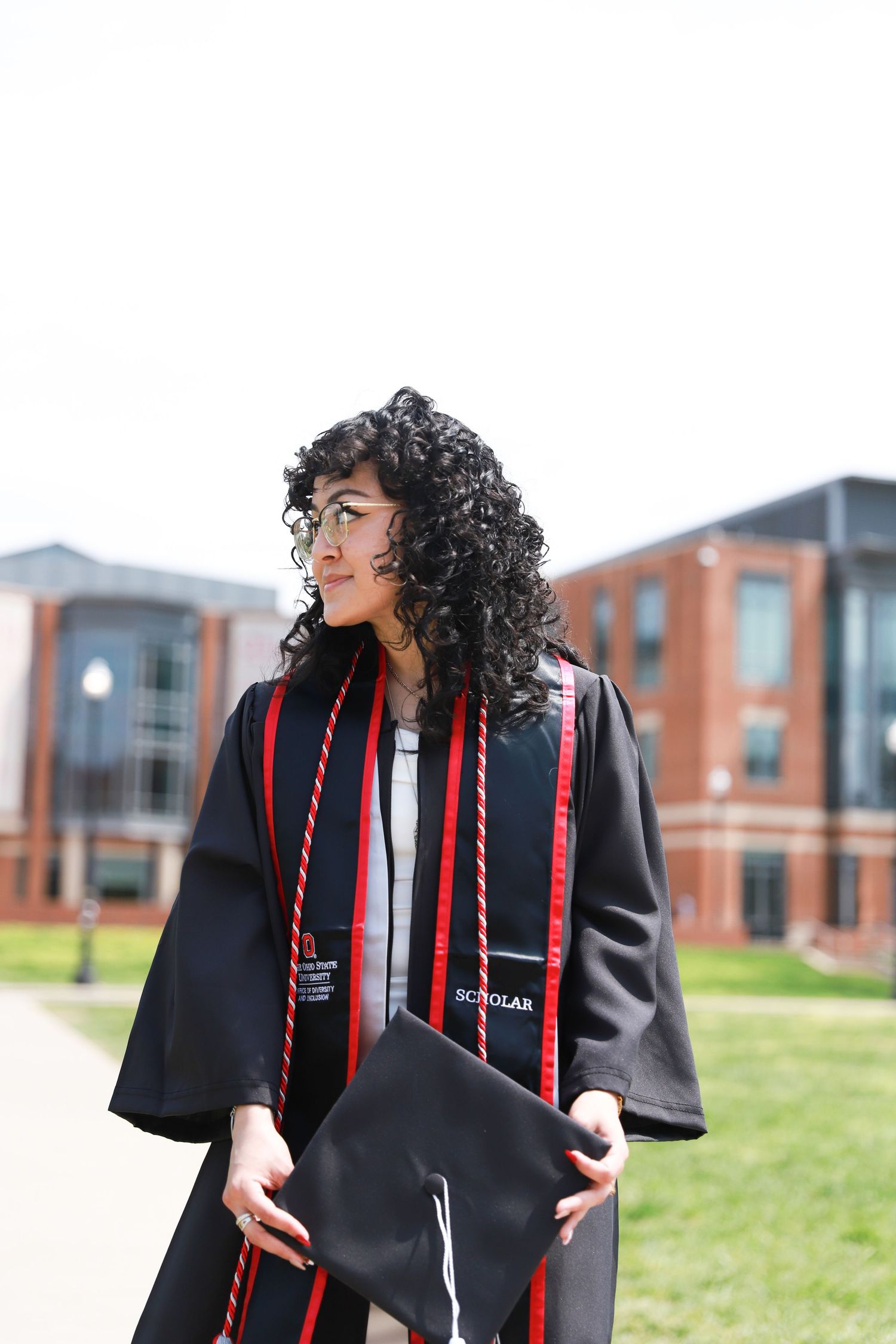 Graduate in black cap and gown with red stole smiles while holding diploma on college campus with brick buildings.
