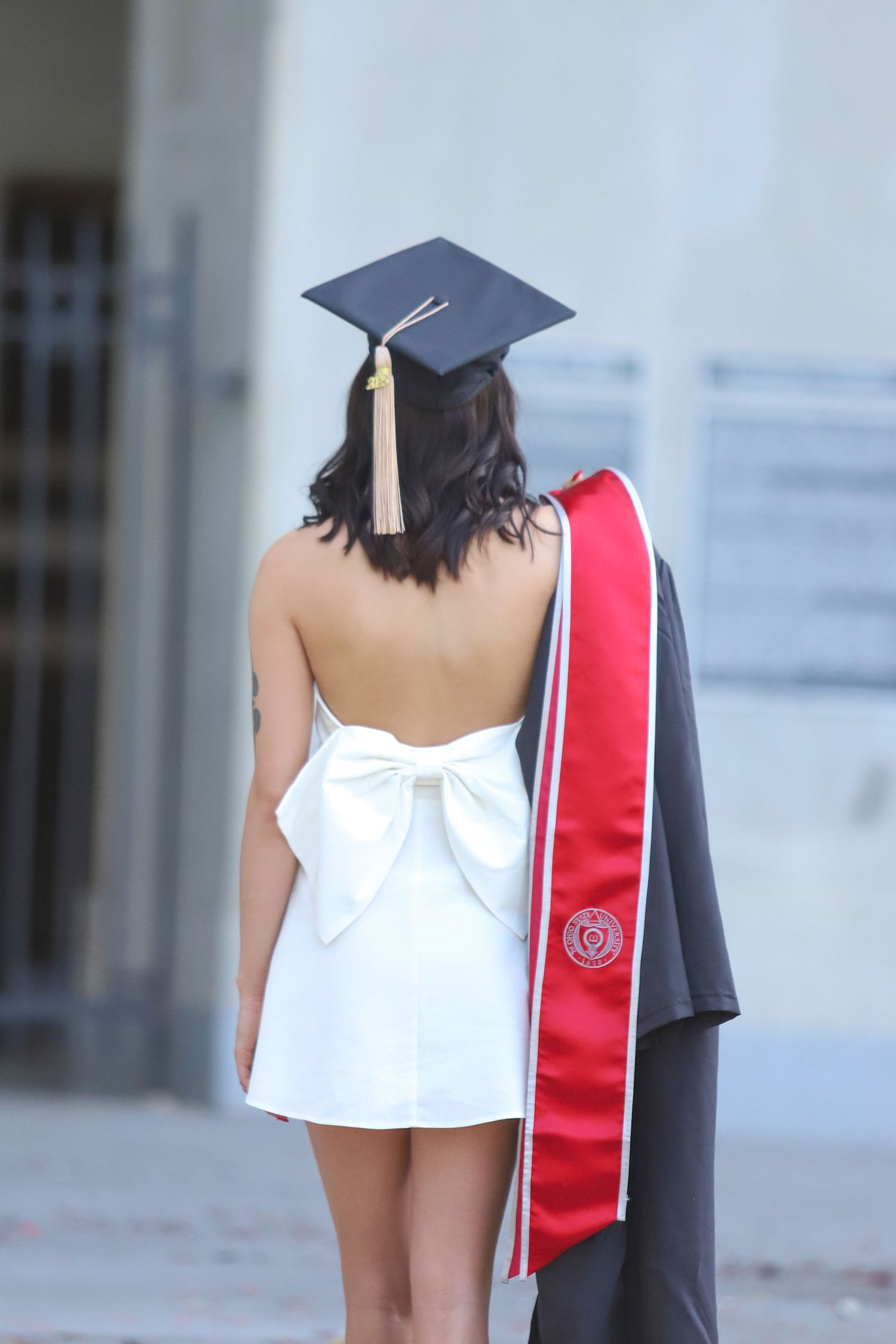 Graduate in white dress and cap poses showing graduation stole and gown from behind.
