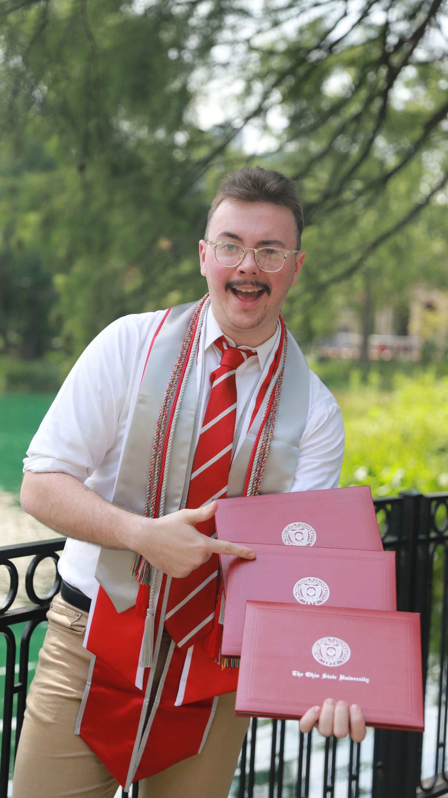 A graduate in suspenders and striped tie holds three red diploma folders while smiling outdoors near a fence.