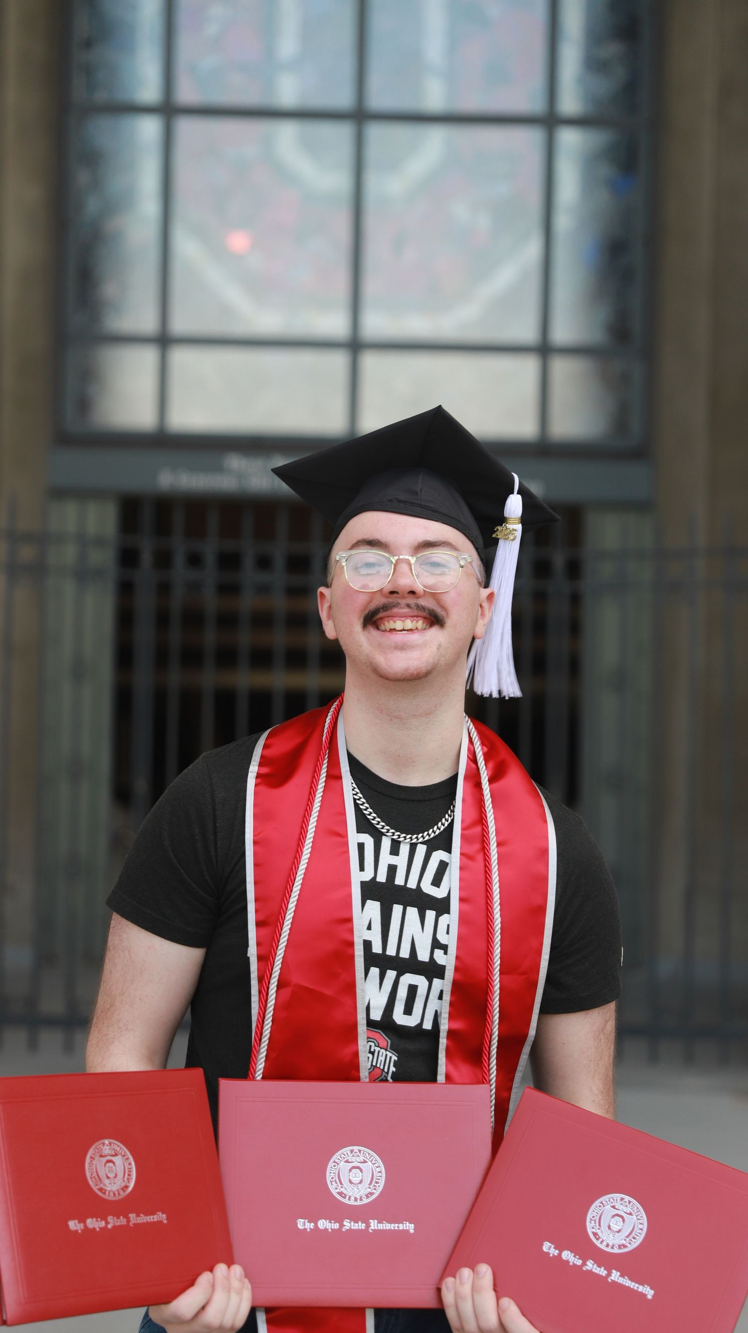 Three graduates in black caps and gowns hold up red diploma folders while posing in front of a glass building.