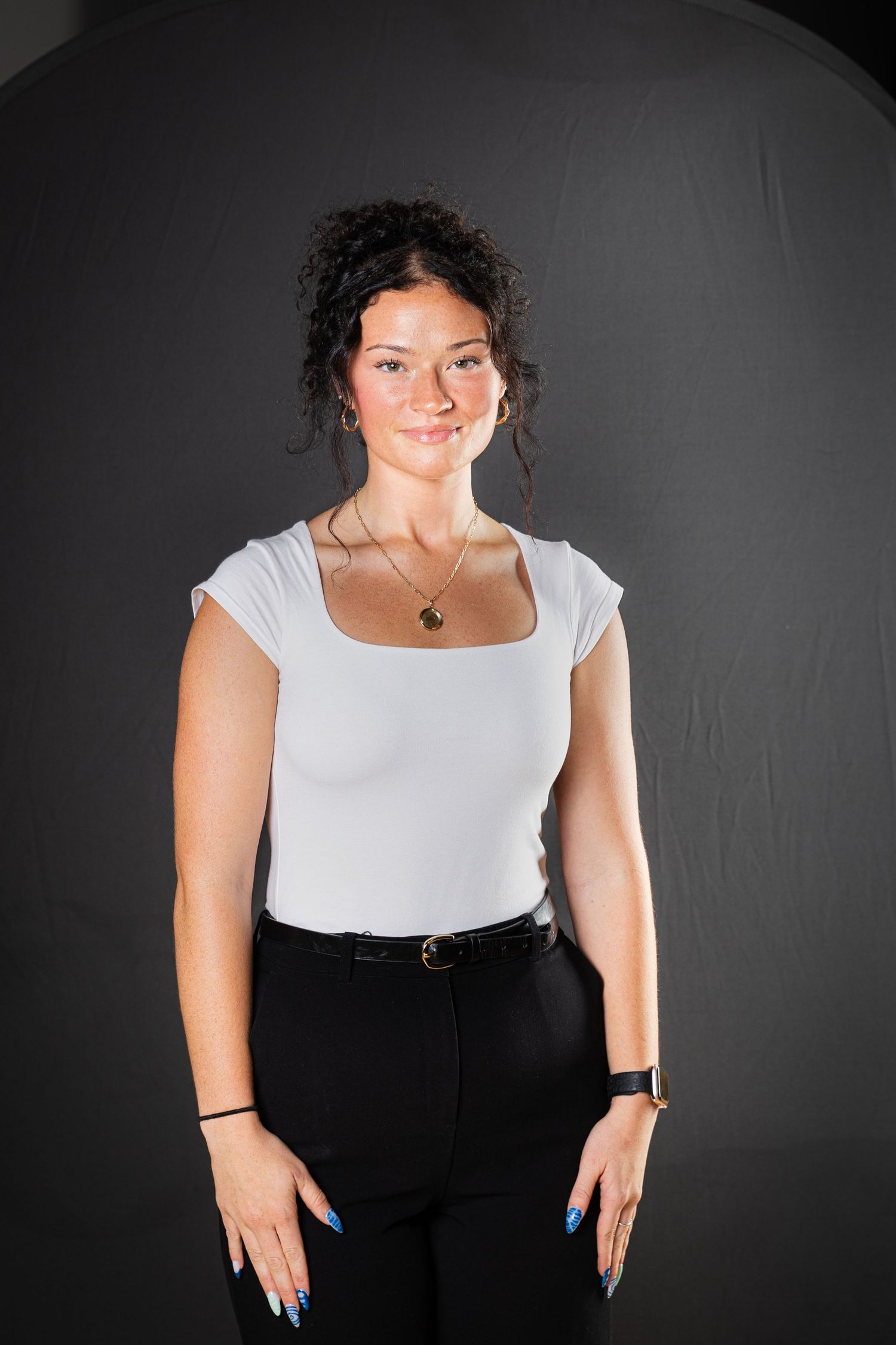 Studio portrait against dark background showing someone in a white top and black pants with bright blue nails.