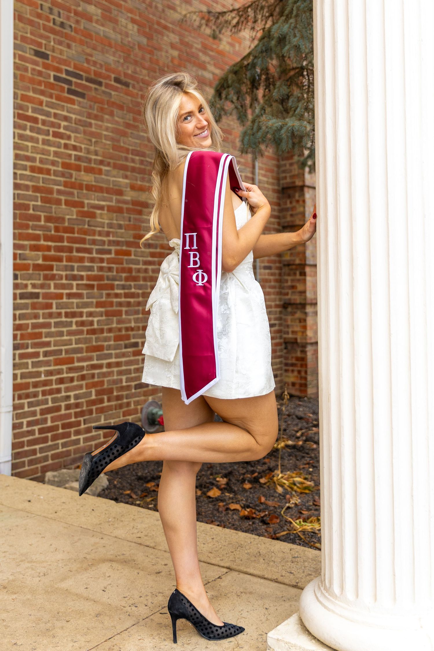 Graduate in white dress and red stole poses next to white column during graduation photoshoot.