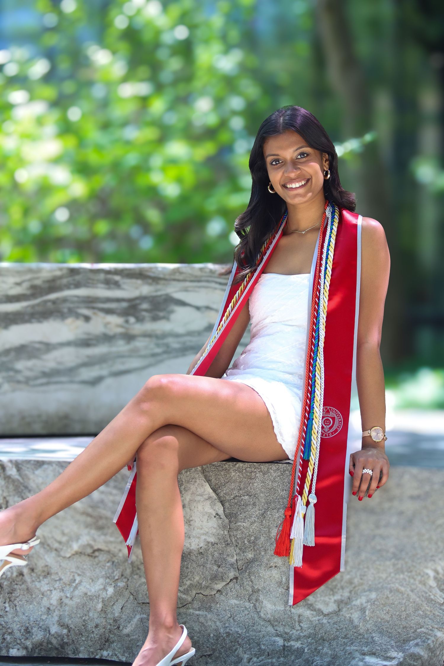 A graduate in a white dress and red graduation stole sits on a rock outdoors during a sunny photoshoot.