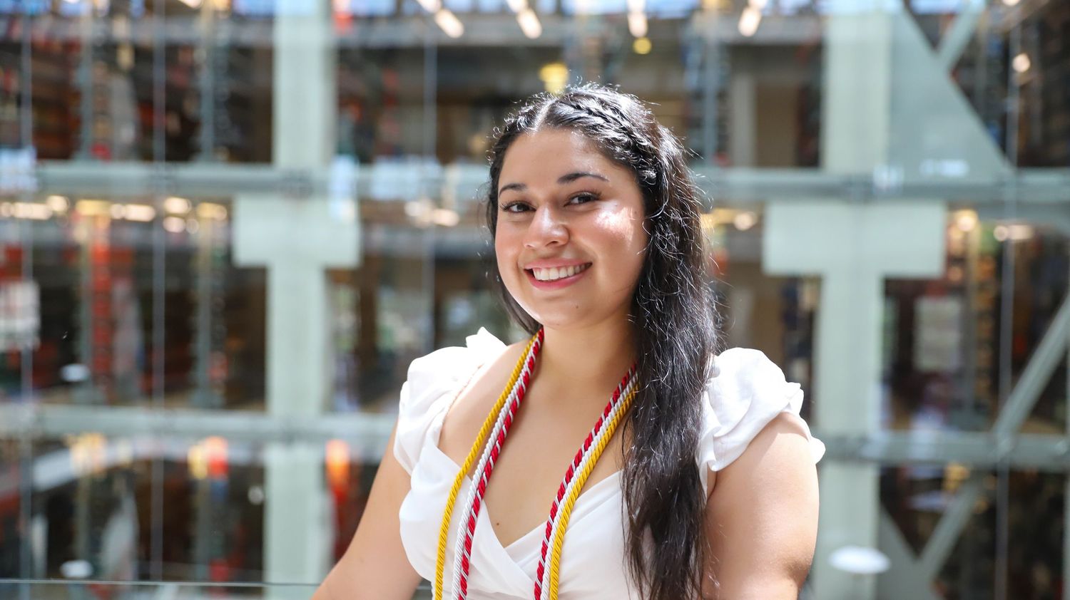 Woman in white dress and bandana poses in front of glass building facade.