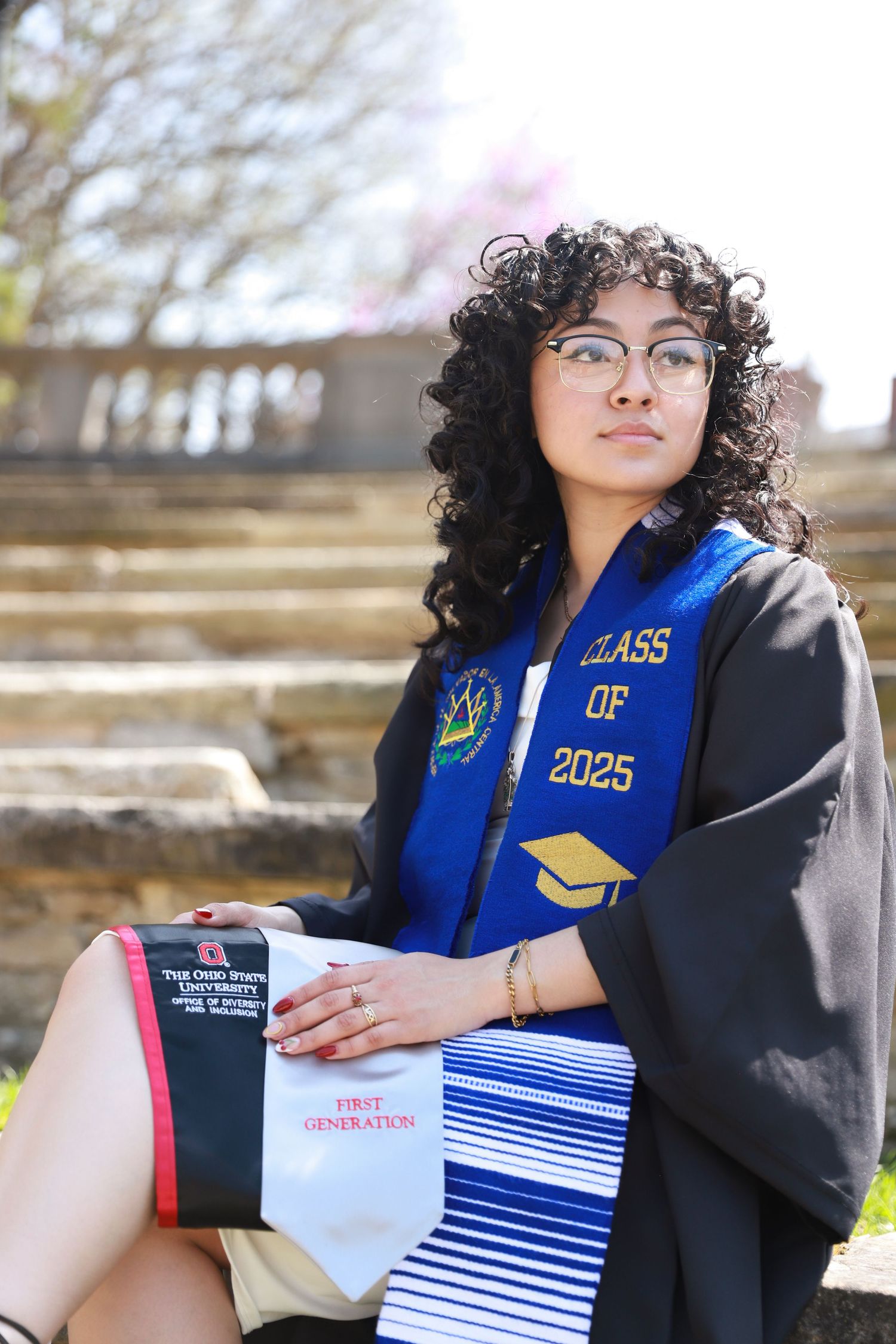 Graduate in blue and black regalia holds diploma while sitting outdoors.