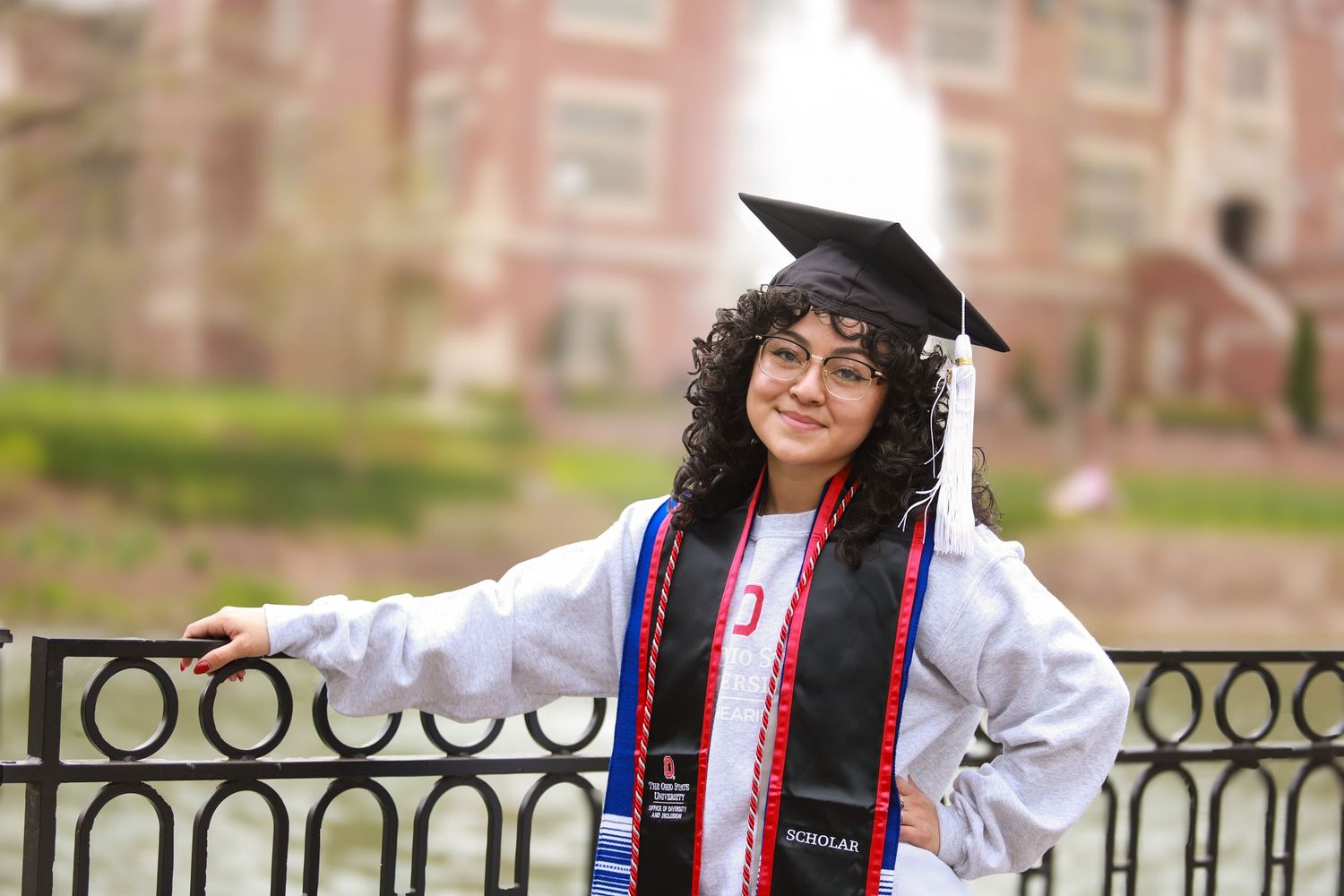 Graduate in cap and gown poses by ornate iron railing during commencement ceremony at brick university building.