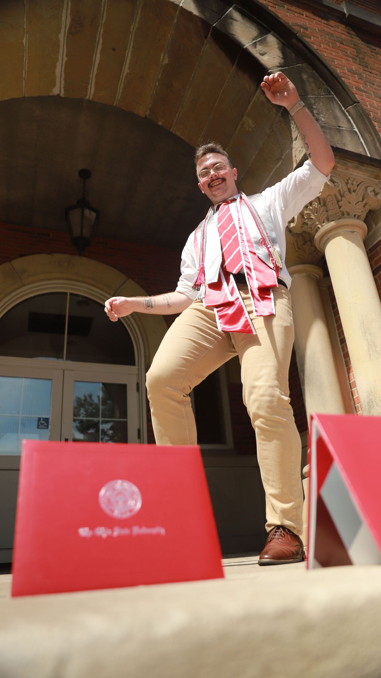 A person in red and white striped shirt and khaki pants dances on a stage with arched columns in the background.