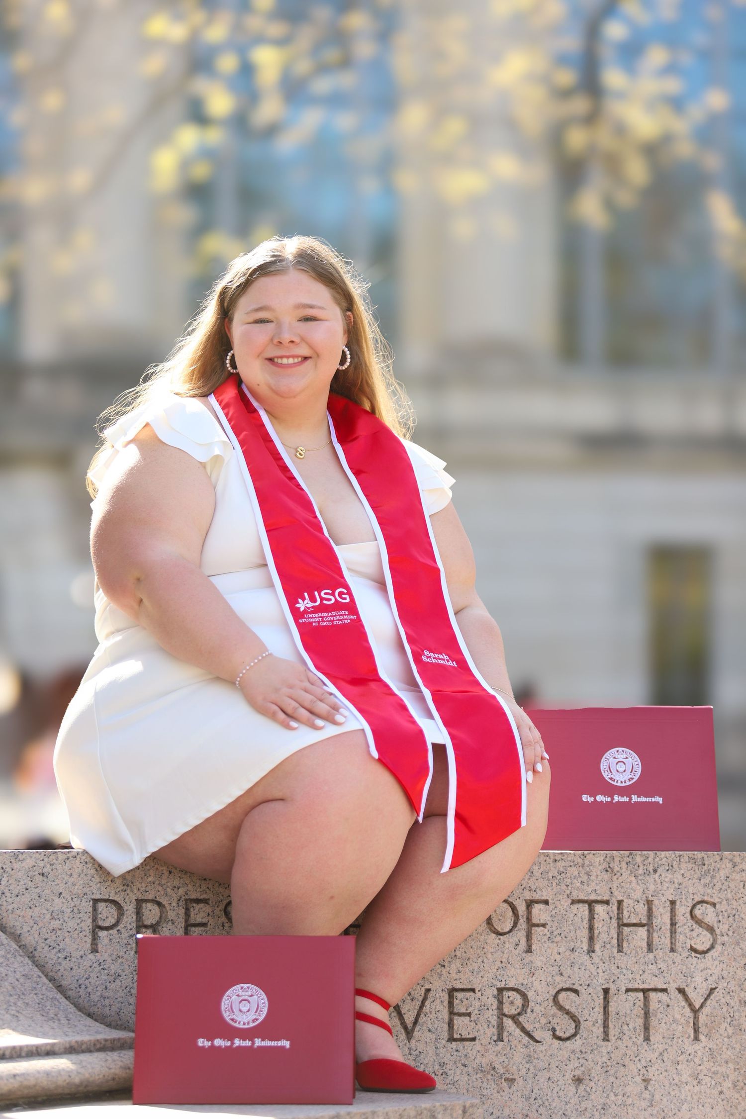 Graduate in white dress and red graduation stole sits on stone wall with diplomas on sunny spring day.