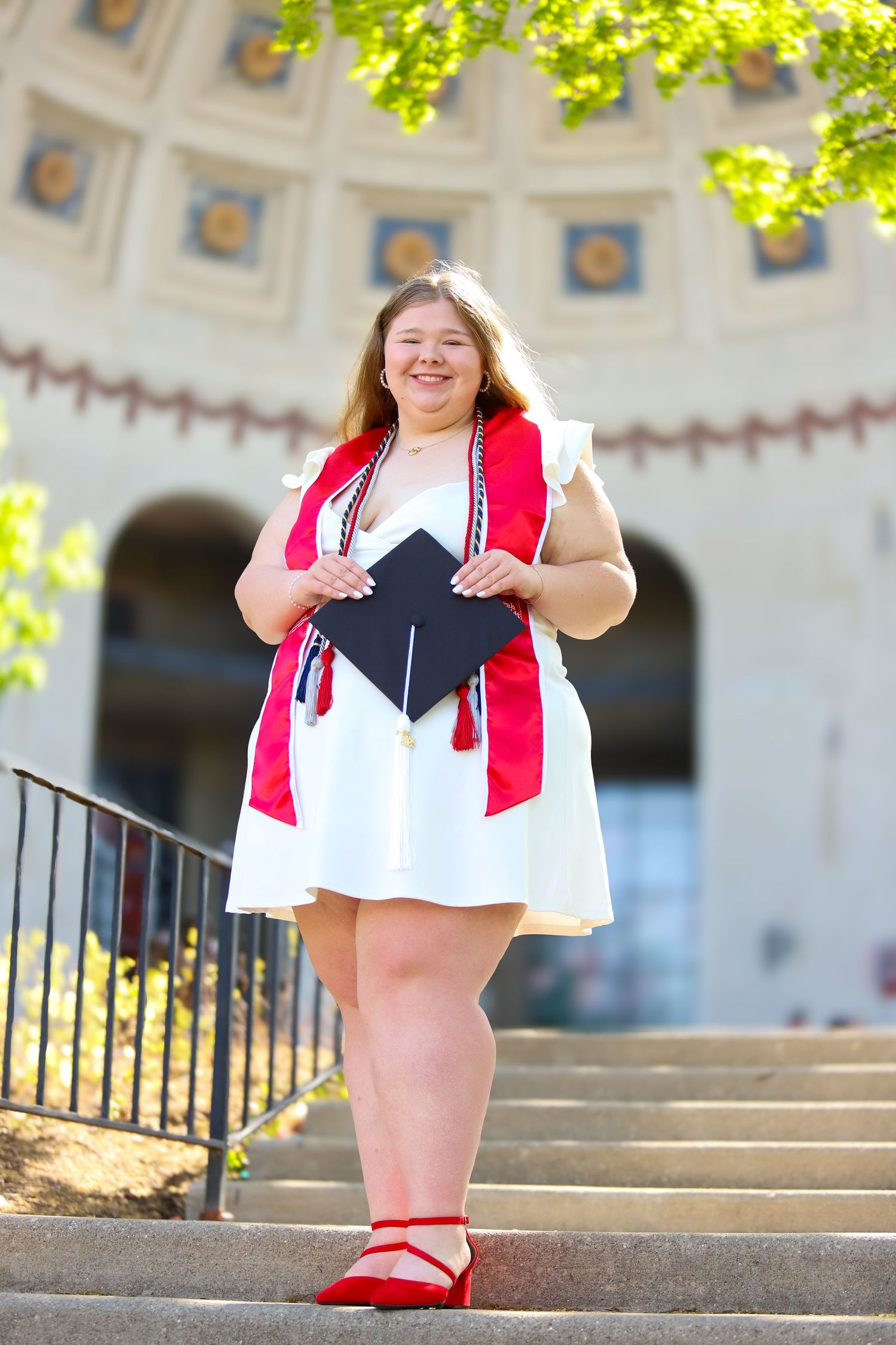 Graduate in cap and gown poses on concrete steps during sunny graduation photoshoot.
