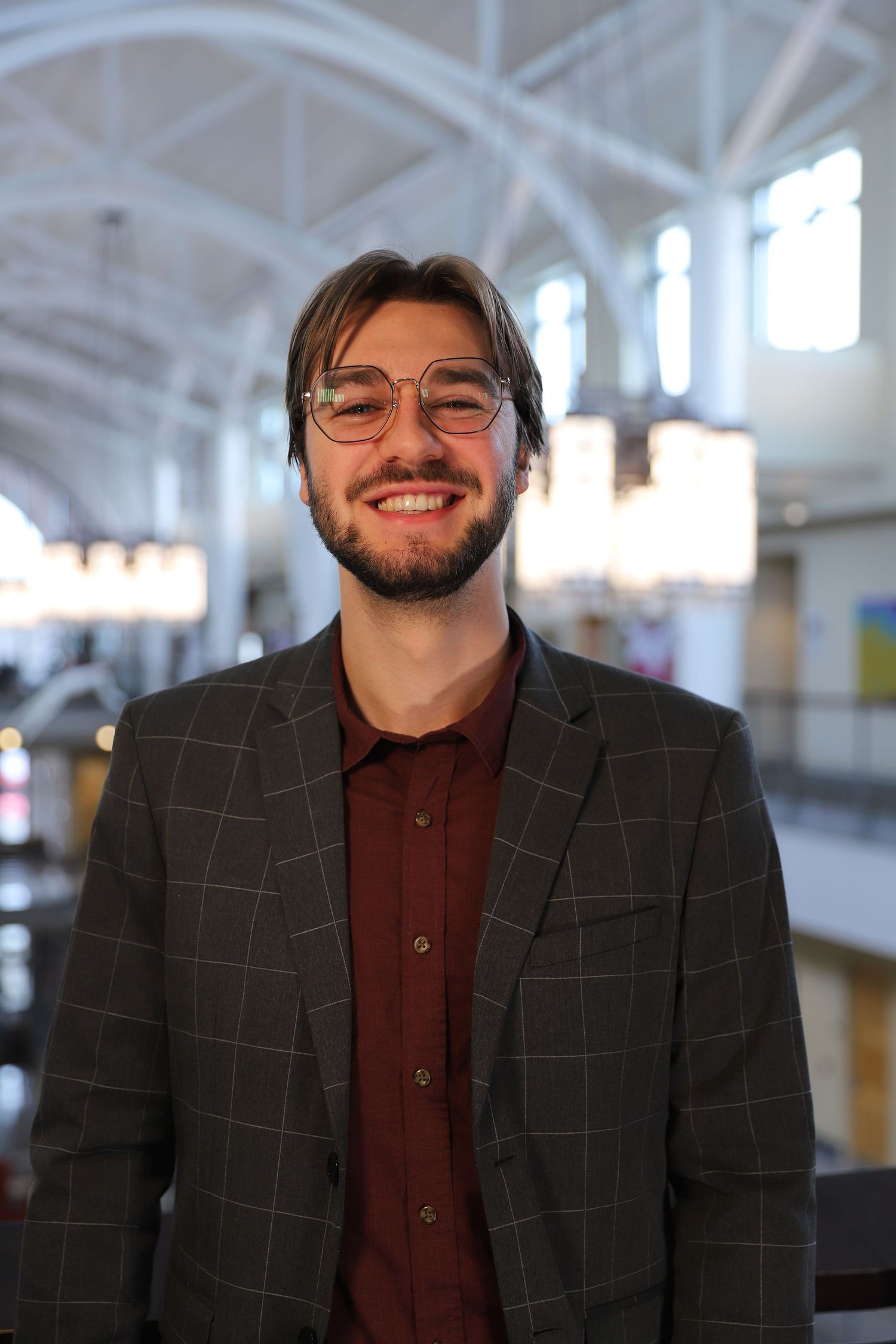 A person wearing glasses and a checkered blazer smiles warmly in a bright indoor setting.