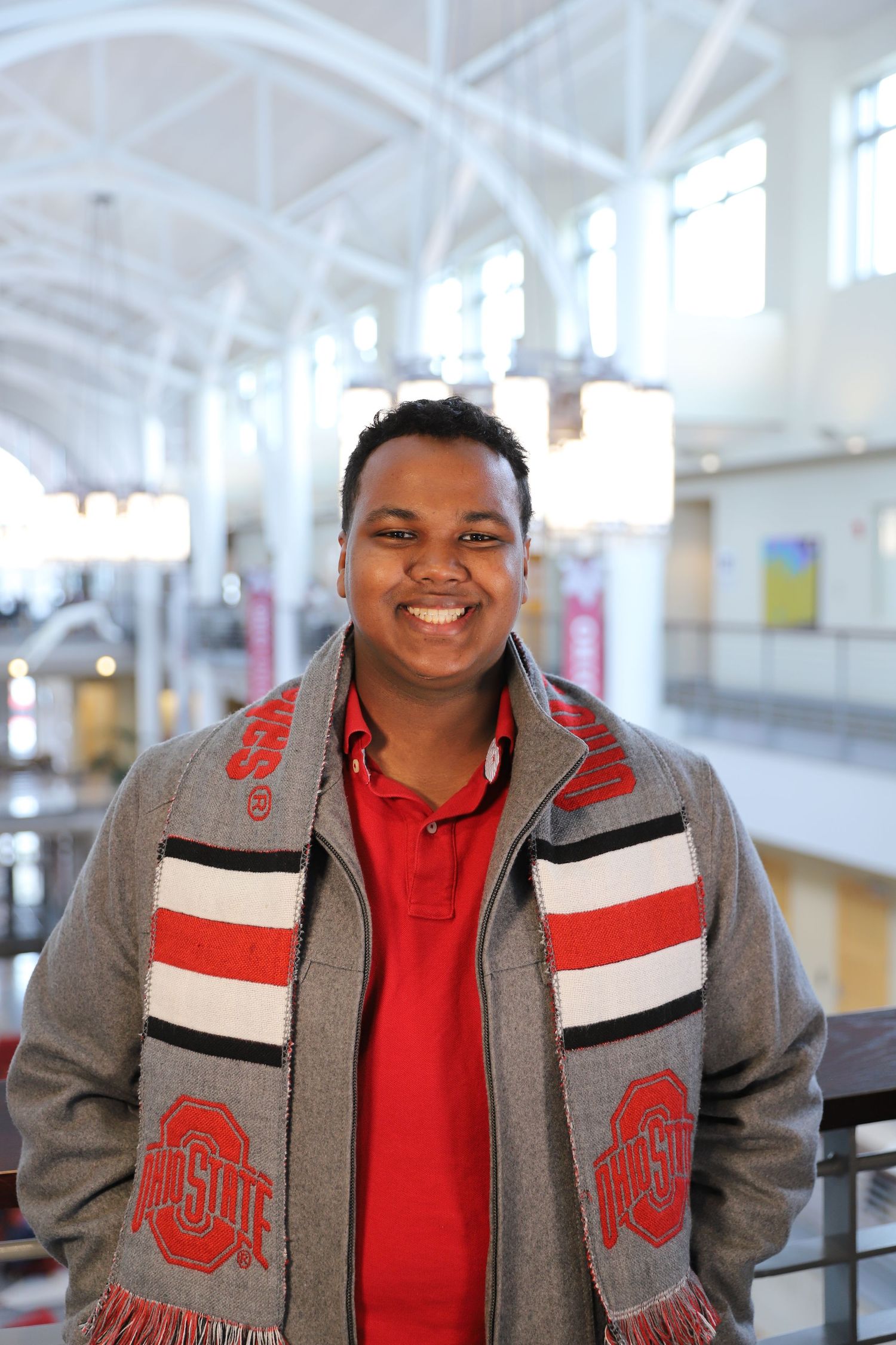 A person wearing a grey and red Ohio State letterman jacket smiles warmly in an indoor setting.