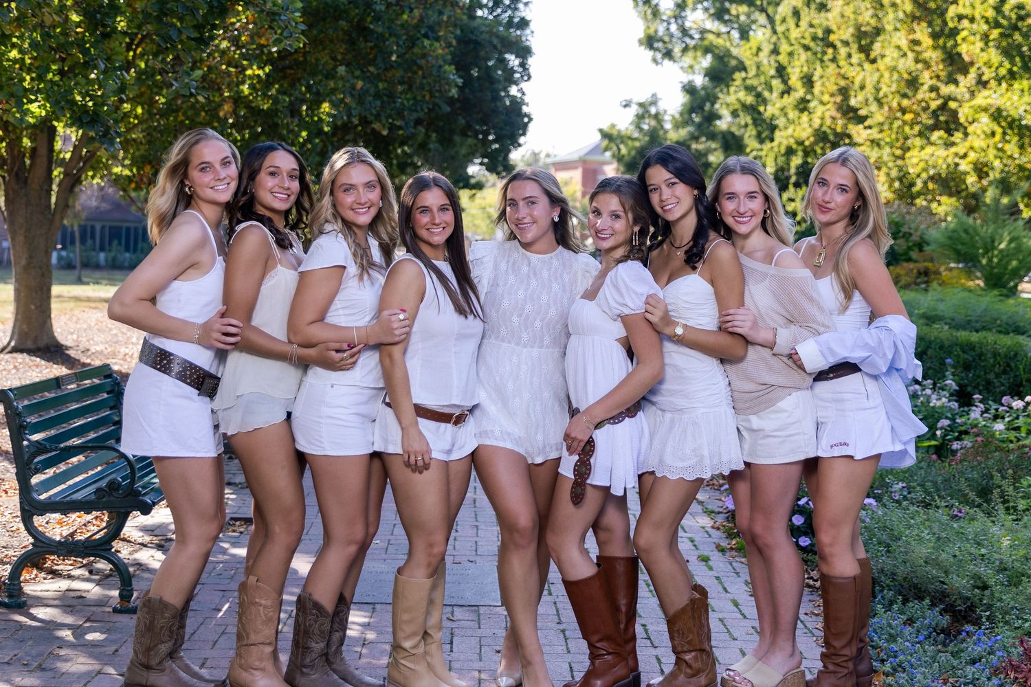 A group in coordinated white outfits poses together outdoors in a garden setting on a sunny day.