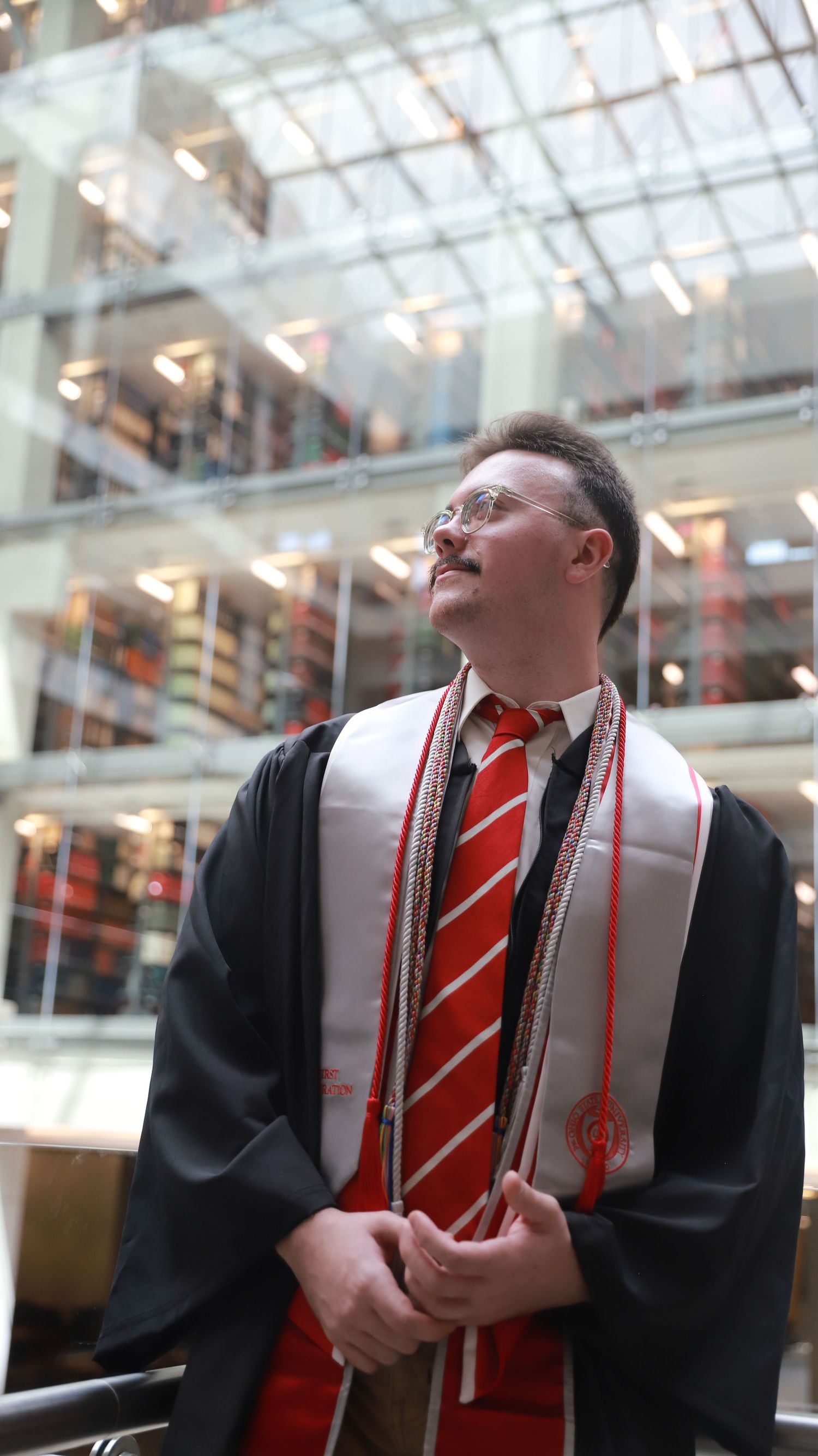 Graduate in black gown and red striped tie laughs joyfully during graduation ceremony in bright atrium.
