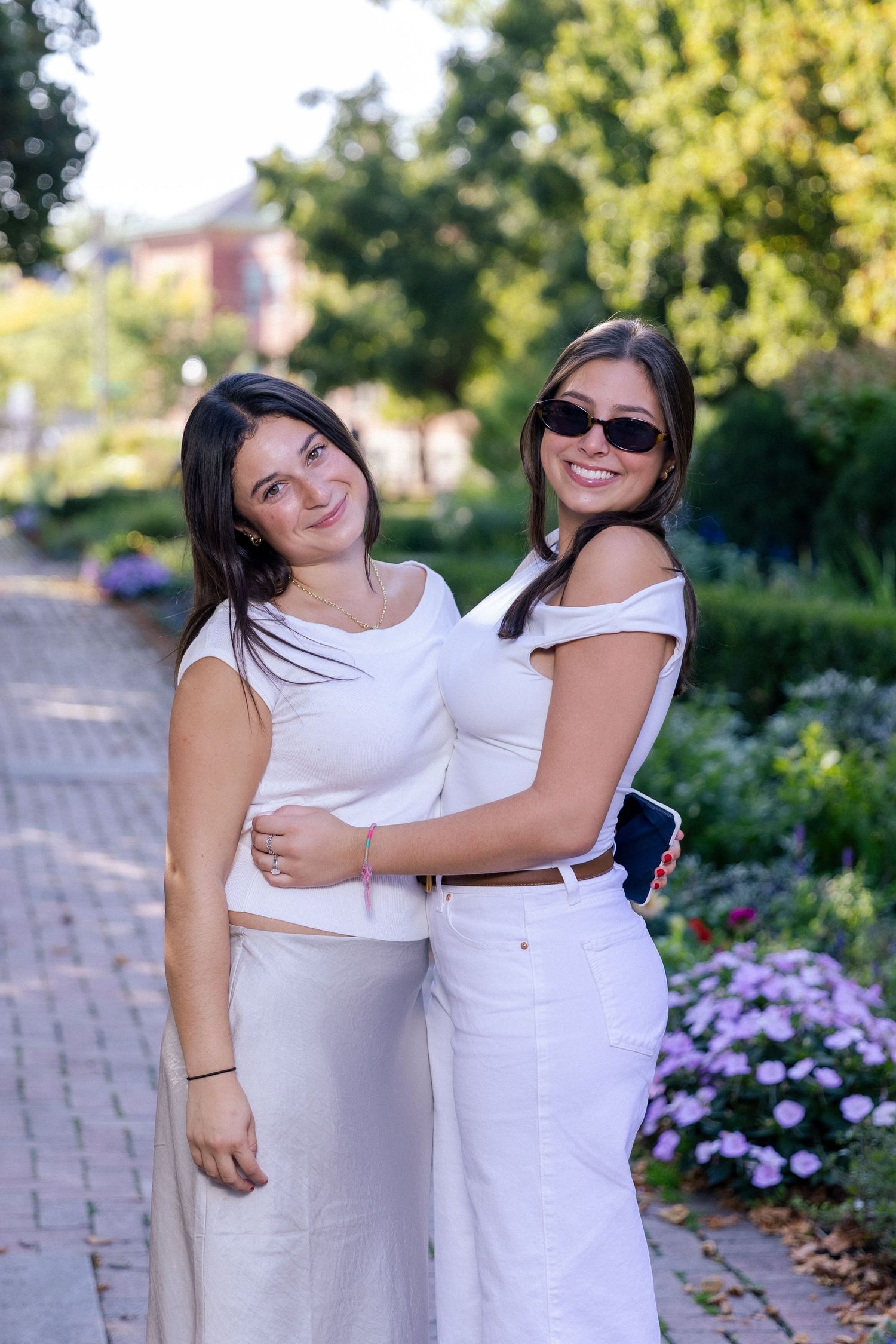 Two people in white dresses embrace while standing on a brick path near blooming purple flowers on a sunny day.