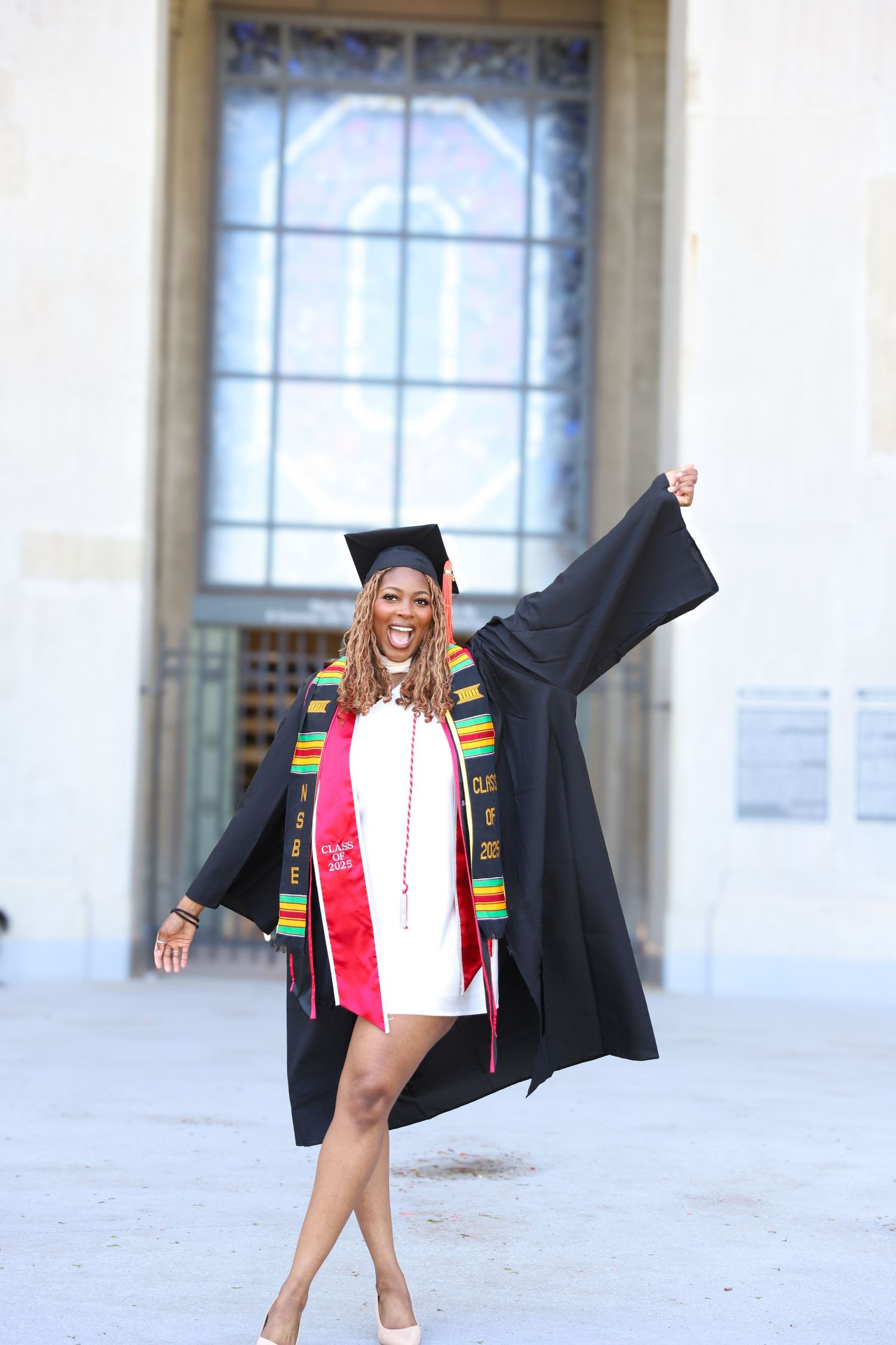 Graduate in black cap and gown with colorful kente stole celebrates joyfully outside building on graduation day.