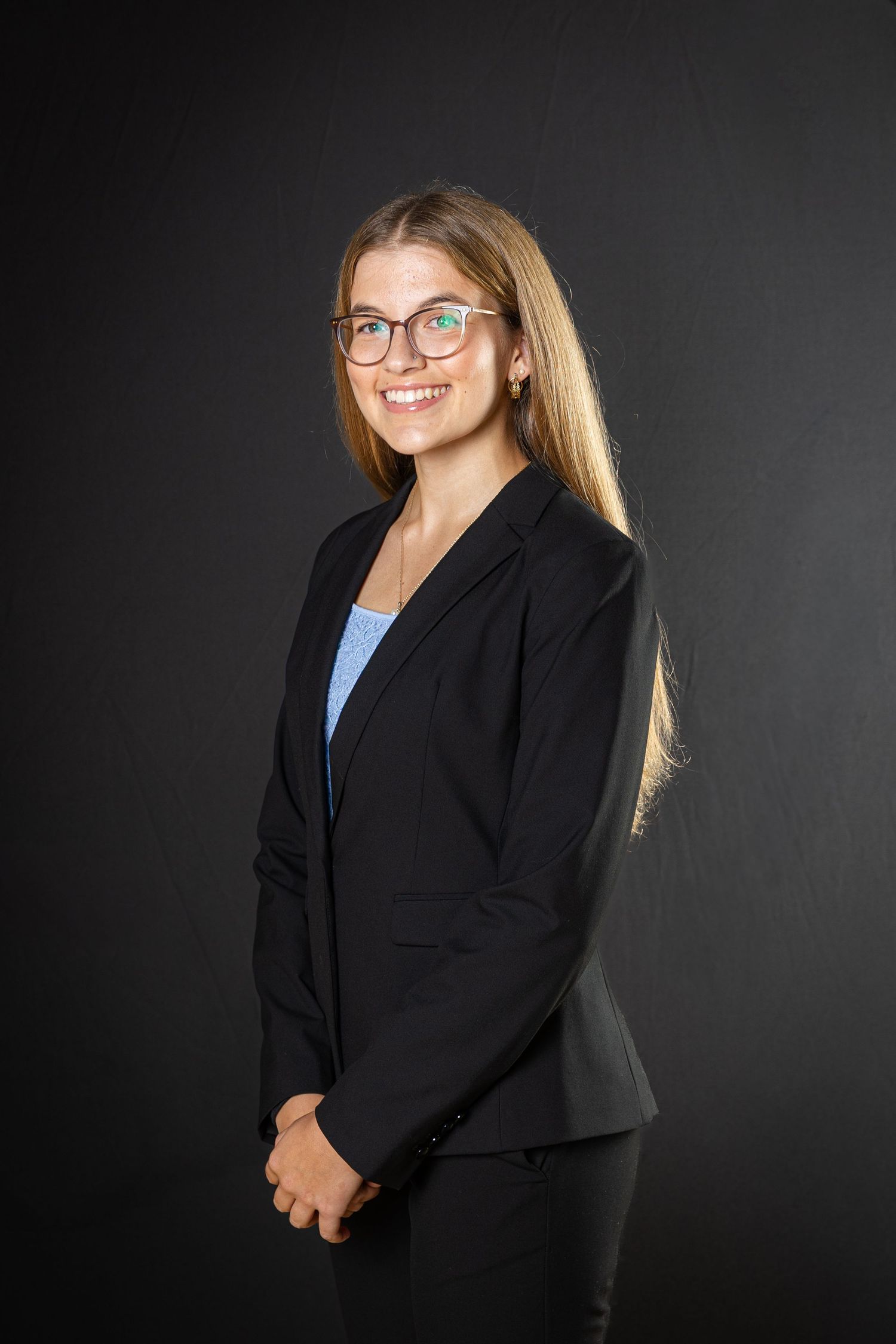 Professional studio portrait of a person in a black blazer and light blue shirt against a dark background.