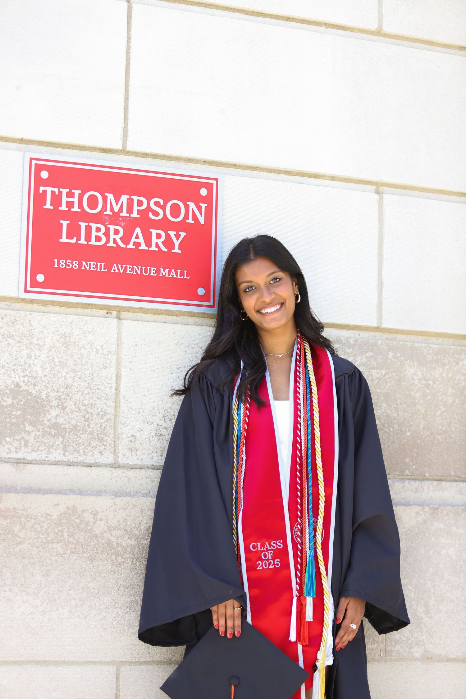 Graduate in navy cap and gown with red stole poses for photos in front of Thompson Library sign.