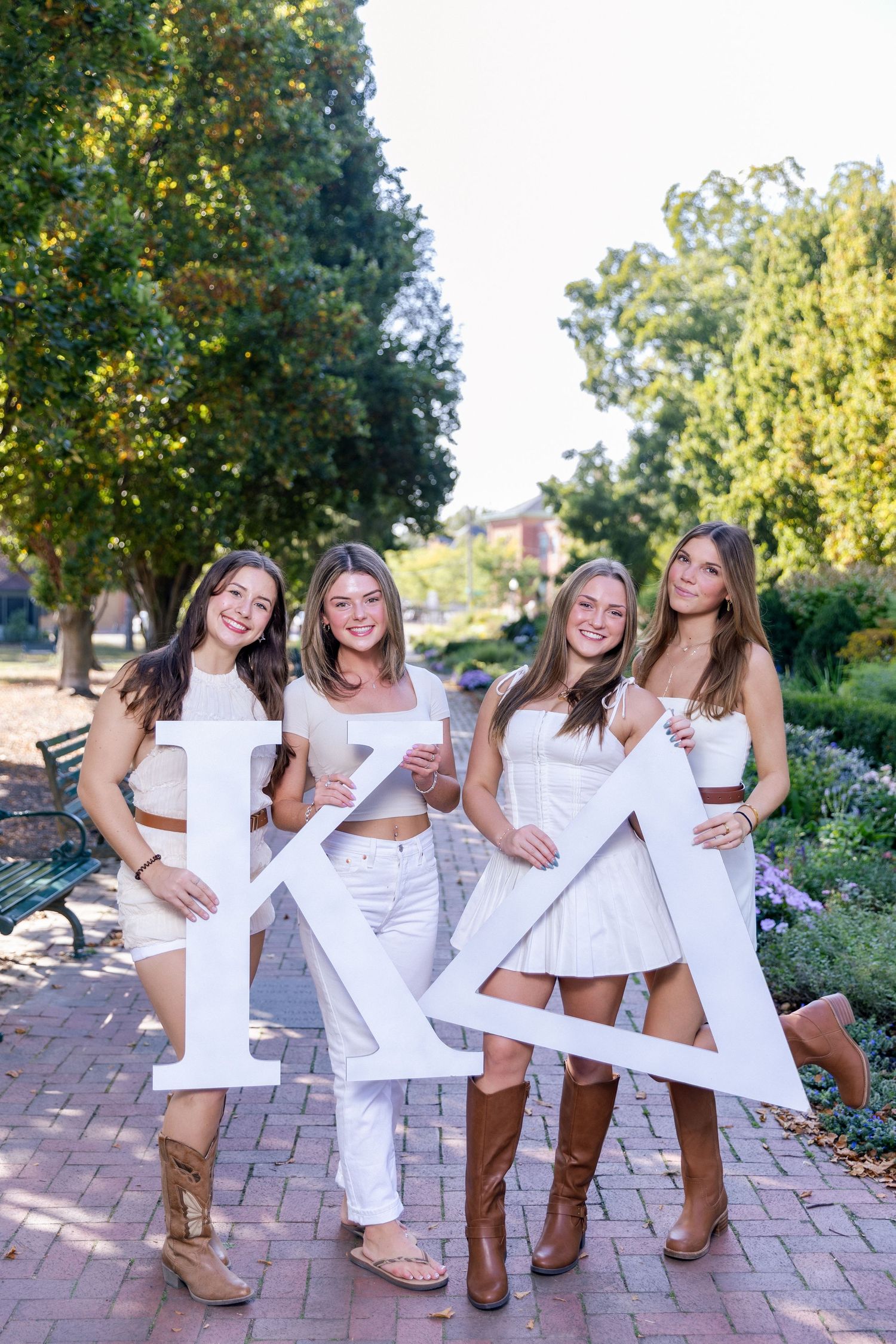 A group wearing matching white outfits and brown boots poses together on a brick patio surrounded by greenery.