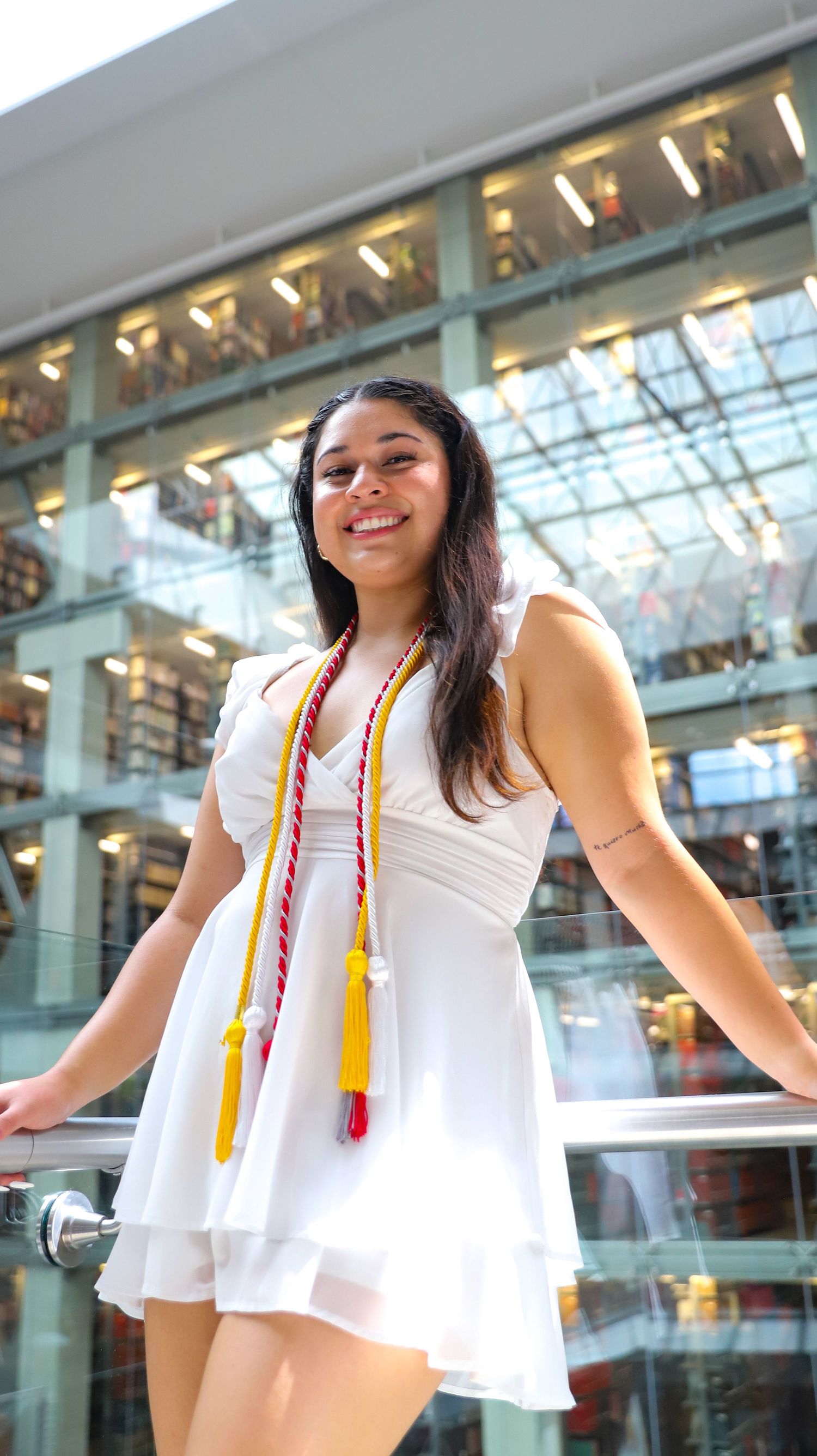 Graduate in white dress with graduation cords poses in modern building with glass ceiling.