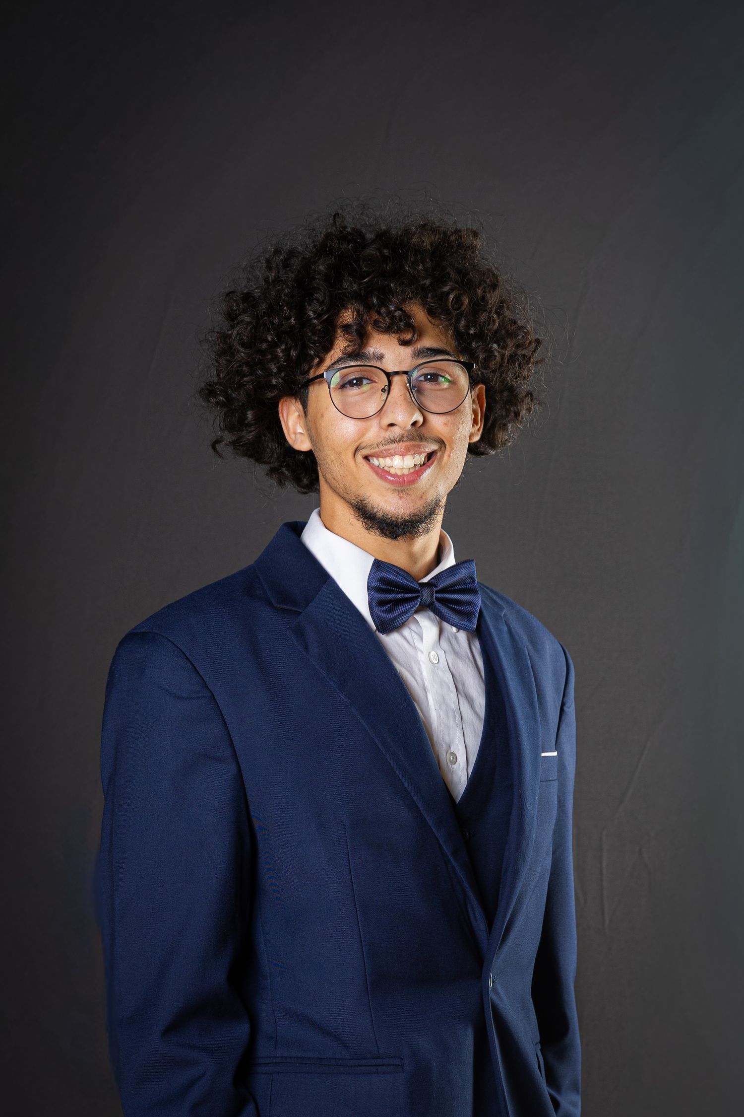 Professional portrait of someone in a navy blue suit with bow tie and glasses against a dark background.
