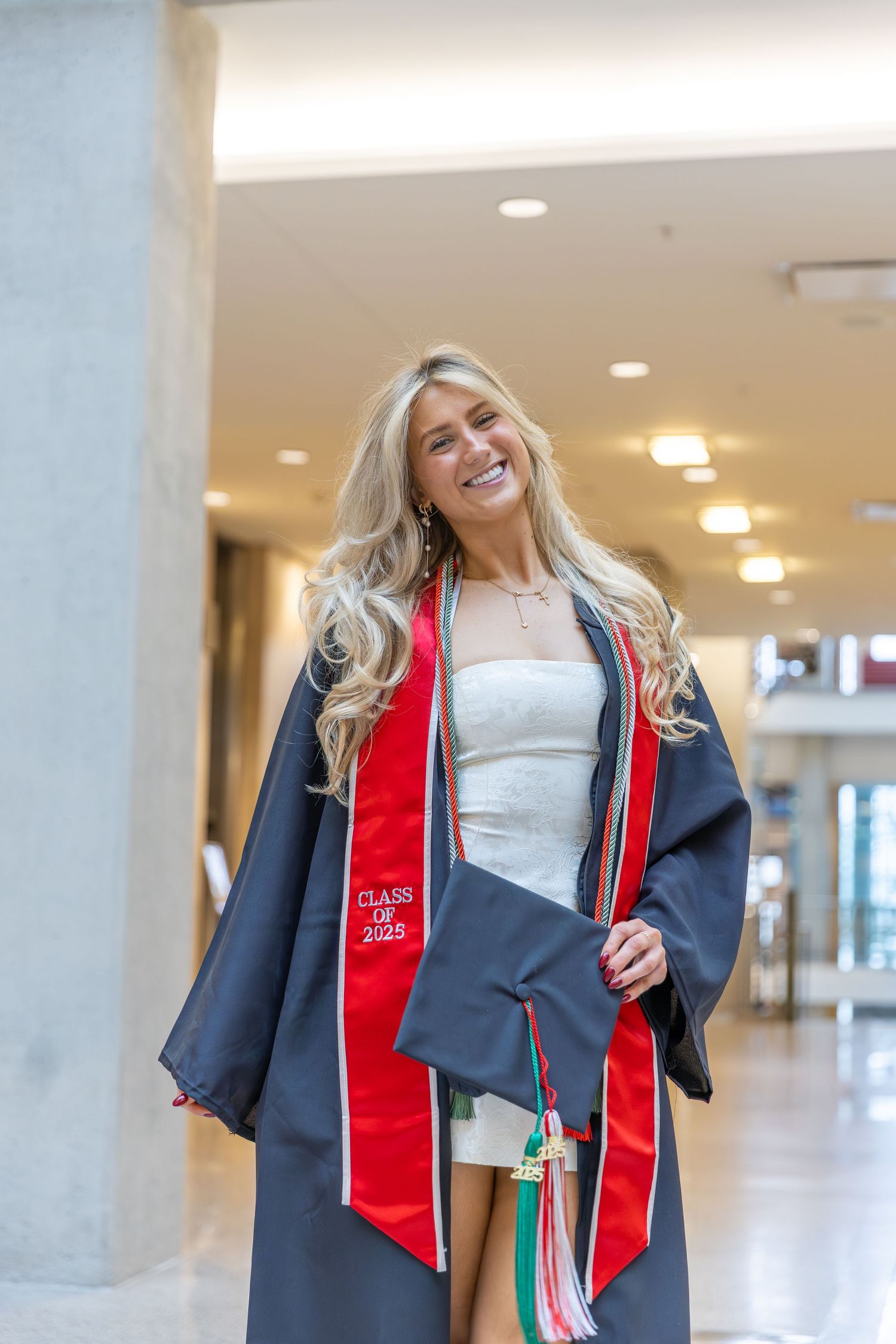 Graduate in cap and gown with red stole walks through a bright indoor hallway.
