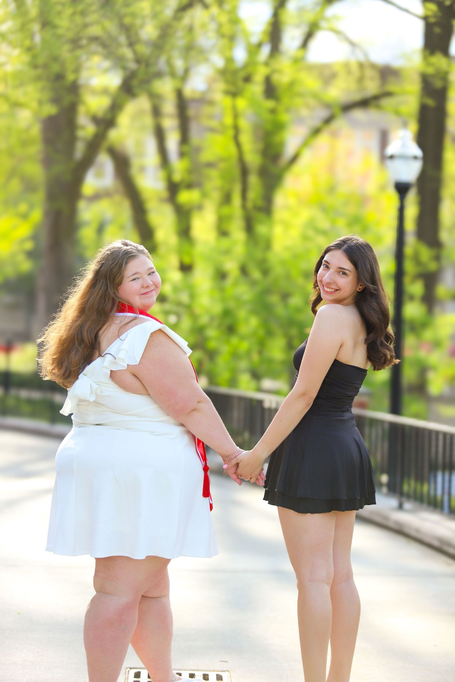 Two dancers in black and white outfits perform graceful movements together on a tree-lined path in a park setting.