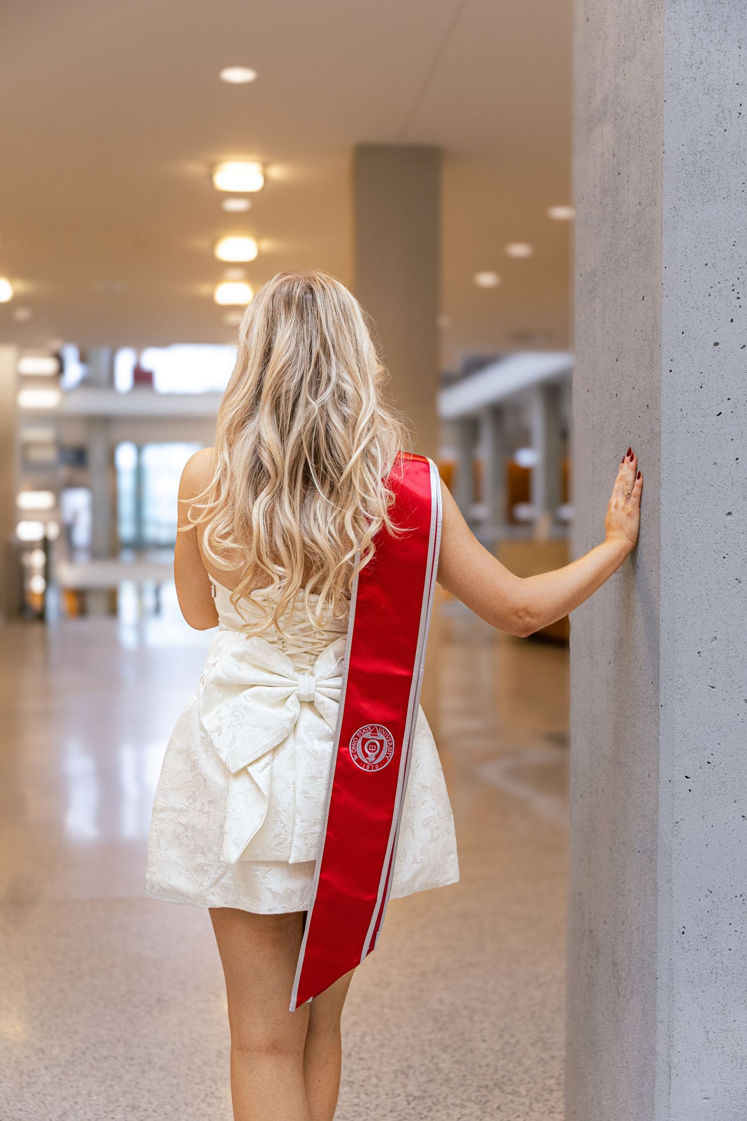 Graduate wearing white dress and red graduation sash stands in hallway with long blonde wavy hair seen from behind.